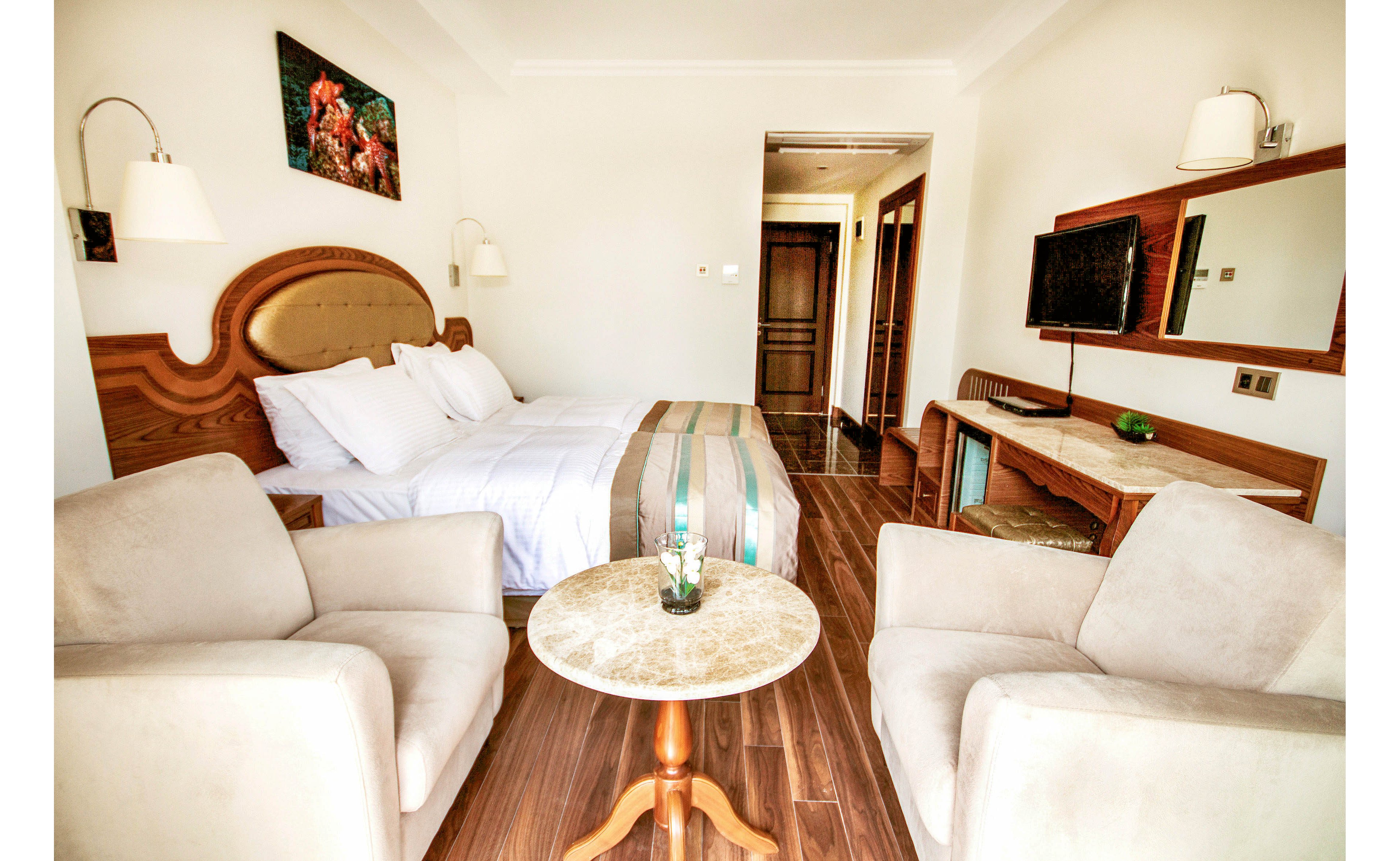 A neatly made double bed with white linens is the focal point of this bright hotel room. Two beige armchairs and a small round table create a cozy seating area on the wooden floor. A wall-mounted flat-screen television, long desk, and large mirror line the opposite wall. Warm lighting and neutral tones give the space a clean, inviting atmosphere.
