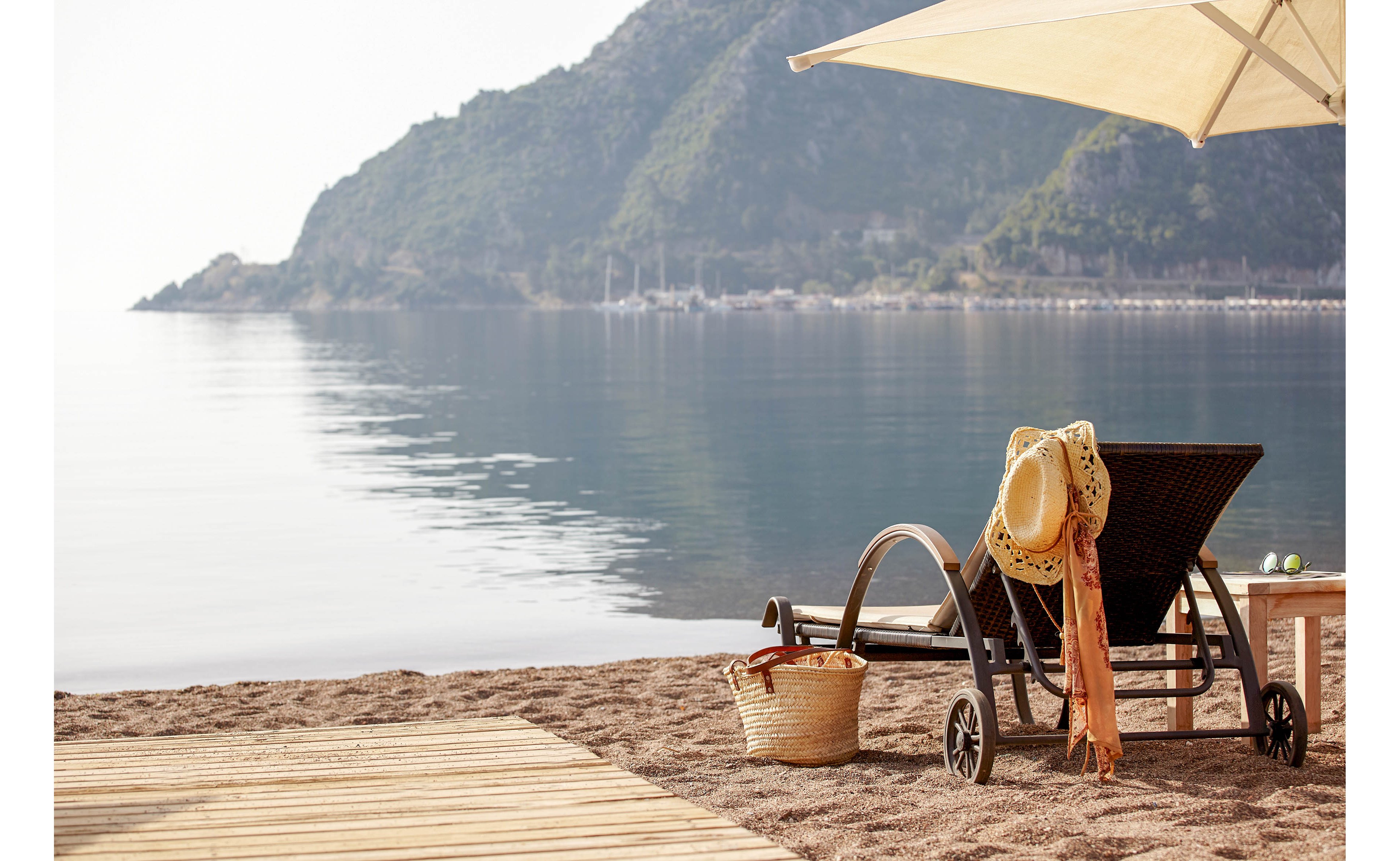 A lounge chair with a straw hat and orange scarf rests on a sandy beach beside a calm sea. A wicker basket and small wooden table sit nearby under a large sun umbrella. In the background, a tree-covered mountain rises along the shoreline, reflected in the still water. The scene is quiet and peaceful, suggesting a relaxing coastal vacation.