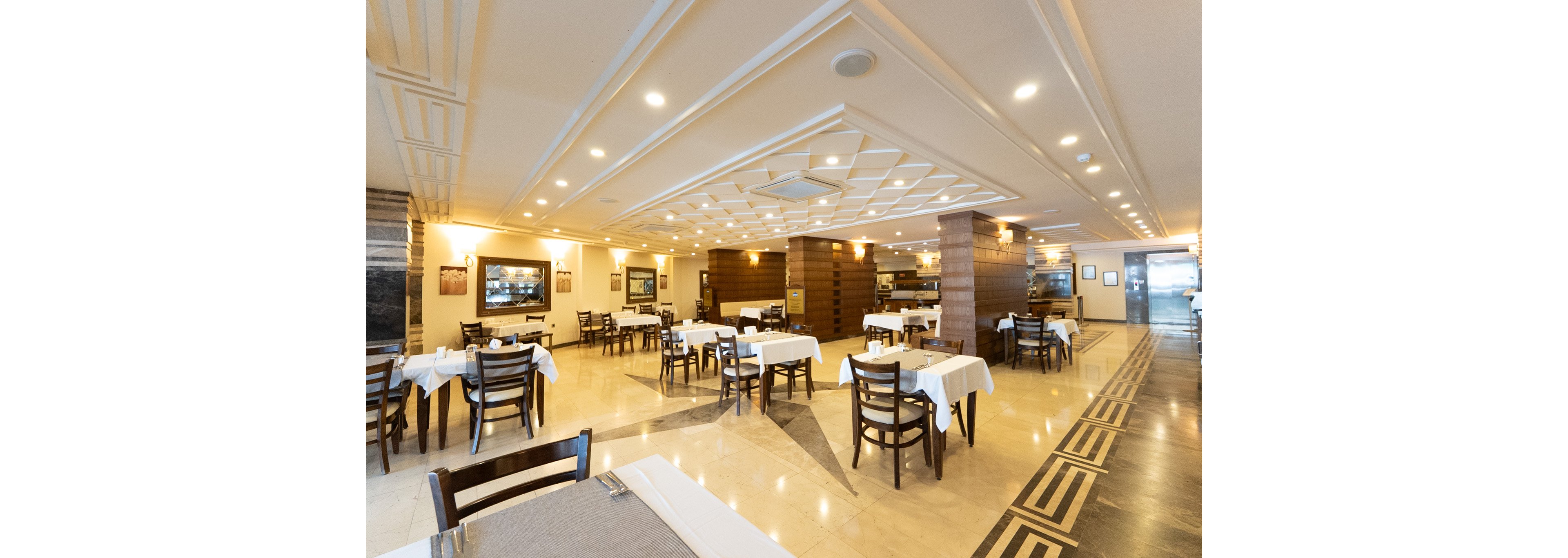 Spacious restaurant dining room with neatly arranged wooden tables and chairs stands empty and ready for guests. White tablecloths and folded napkins create a clean, formal look across the polished floor. Warm ceiling lights and recessed lighting highlight the decorative ceiling panels and wooden columns. Framed artwork and a long service counter are visible in the background, adding depth to the inviting interior.