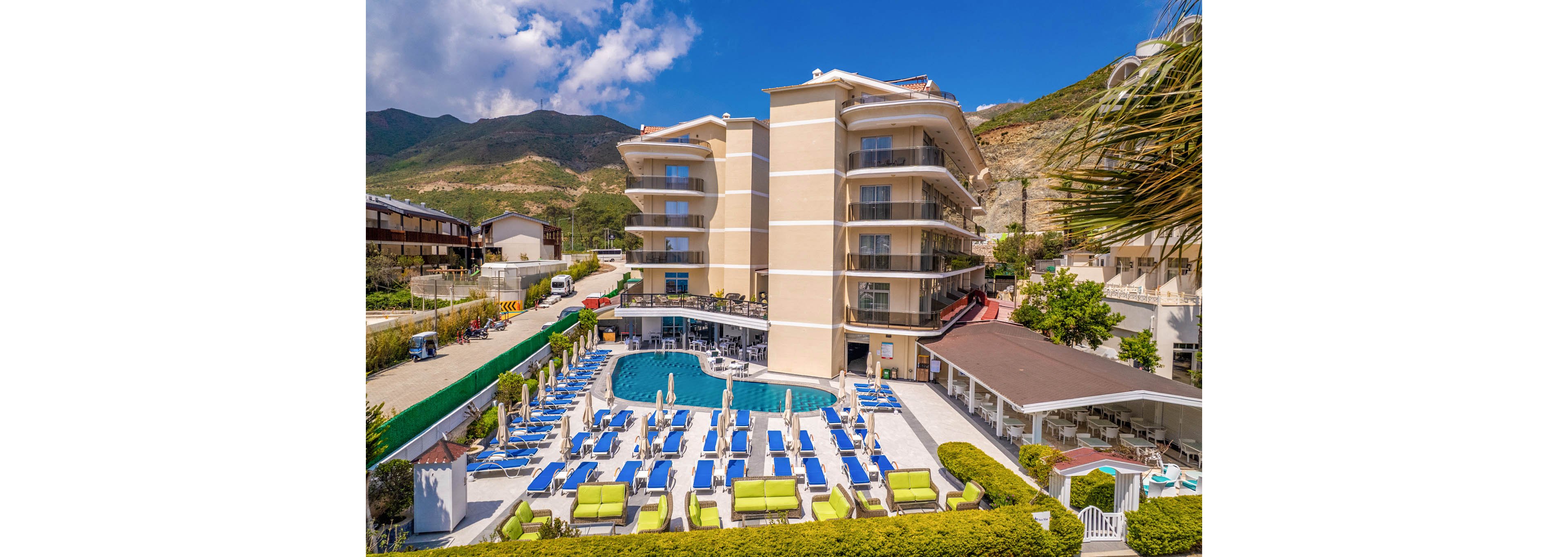 A modern multi-story resort hotel features a large outdoor swimming pool surrounded by numerous blue and white sun loungers. The building is set against a backdrop of green hills and a partly cloudy blue sky. Guests can relax on poolside seating areas, including cushioned sofas and shaded terraces. The scene suggests a sunny vacation destination with well-maintained landscaping and a calm atmosphere.