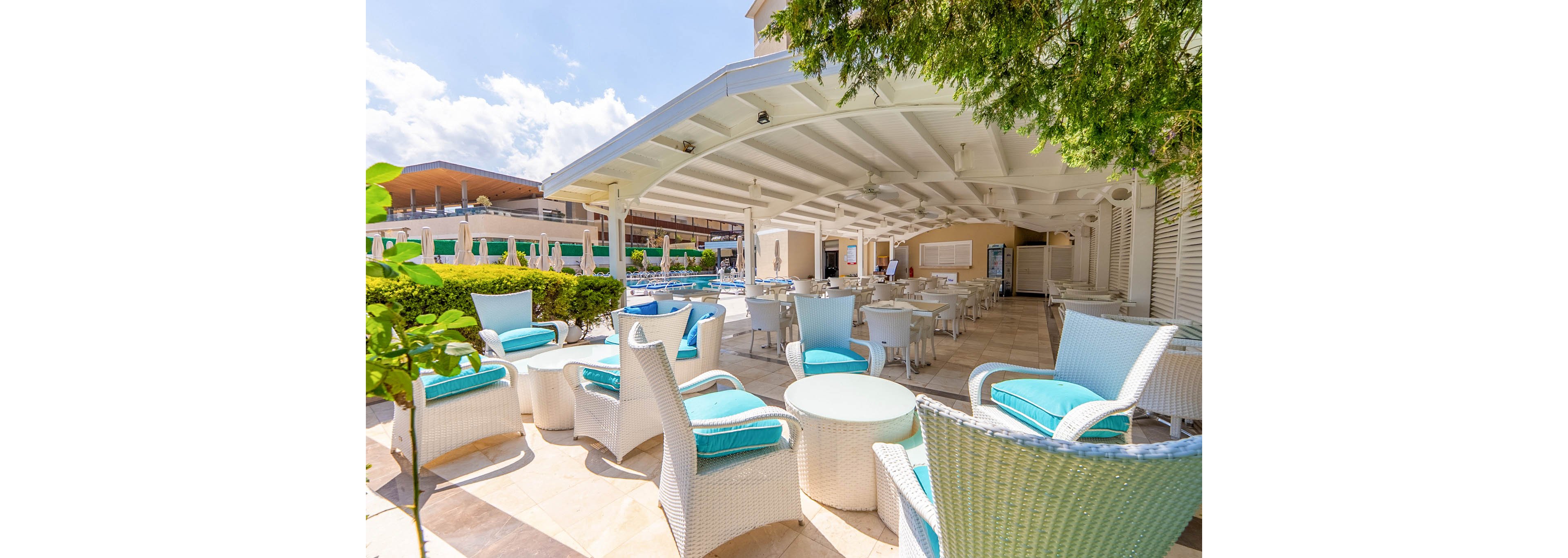 An open air resort lounge features white wicker furniture with bright turquoise cushions arranged around small round tables. The covered patio area opens onto a poolside setting with additional seating and sunlit walkways. Ceiling fans and soft natural light create a relaxed, breezy atmosphere. Lush greenery and modern resort buildings frame the scene, suggesting a comfortable vacation or leisure environment.