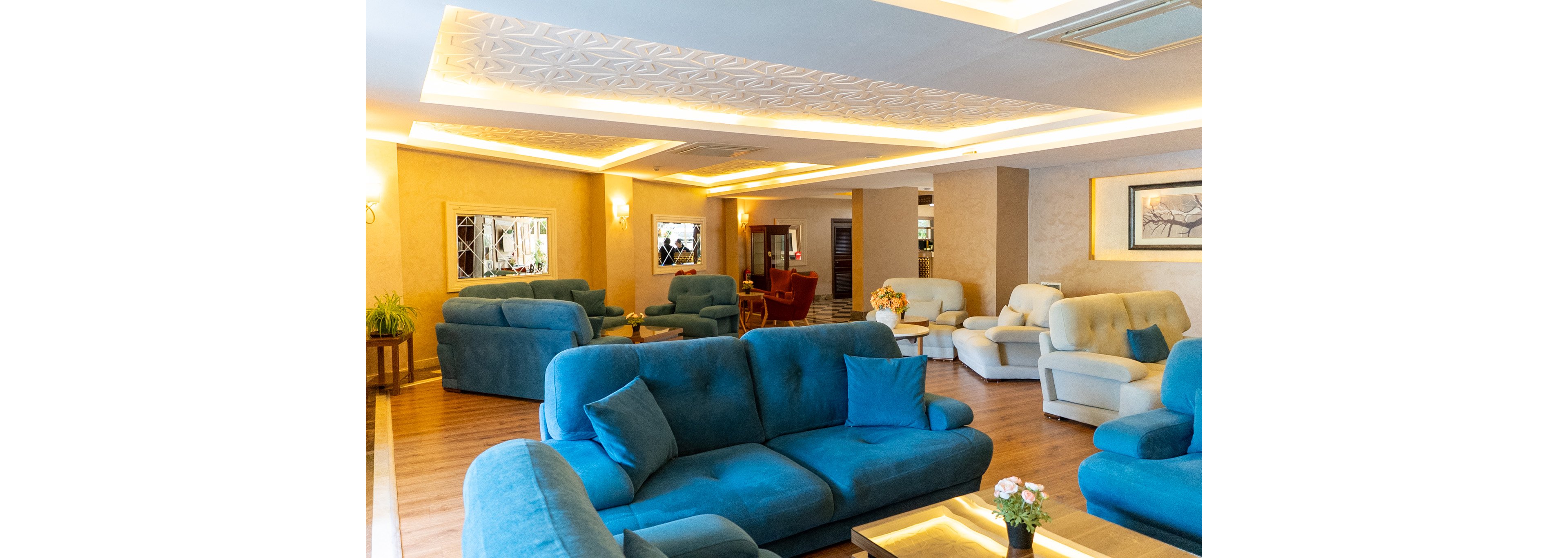 A spacious lounge area features plush blue and beige sofas arranged for relaxed seating. Warm ambient lighting from ceiling fixtures and wall sconces creates a cozy atmosphere. Wooden flooring and decorative ceiling details add a touch of elegance. Small tables with flower arrangements and framed artwork on the walls complete the contemporary interior design.