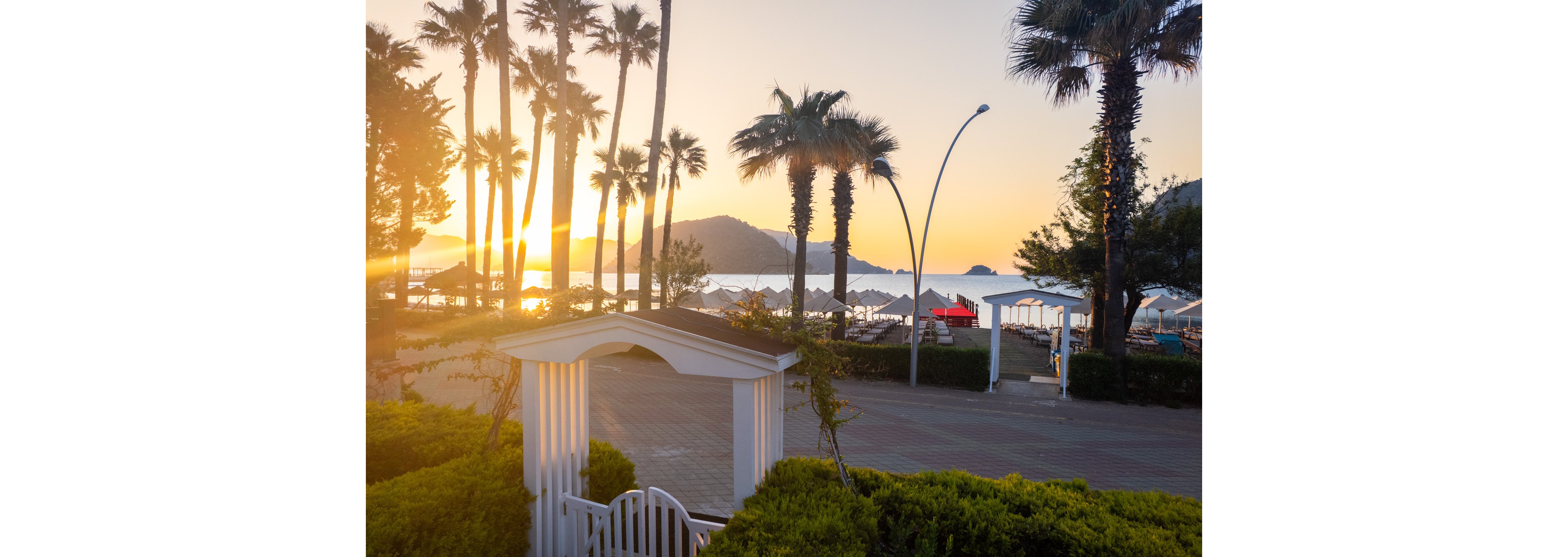 A tranquil beachfront resort is captured at sunrise with the sun glowing low over the sea. Tall palm trees frame the view, casting long shadows across the promenade and sandy beach. White beach umbrellas and lounge chairs line the shore, while a white garden gate and manicured hedges appear in the foreground. Distant hills and small islands are silhouetted against the warm, golden sky, creating a peaceful holiday atmosphere.