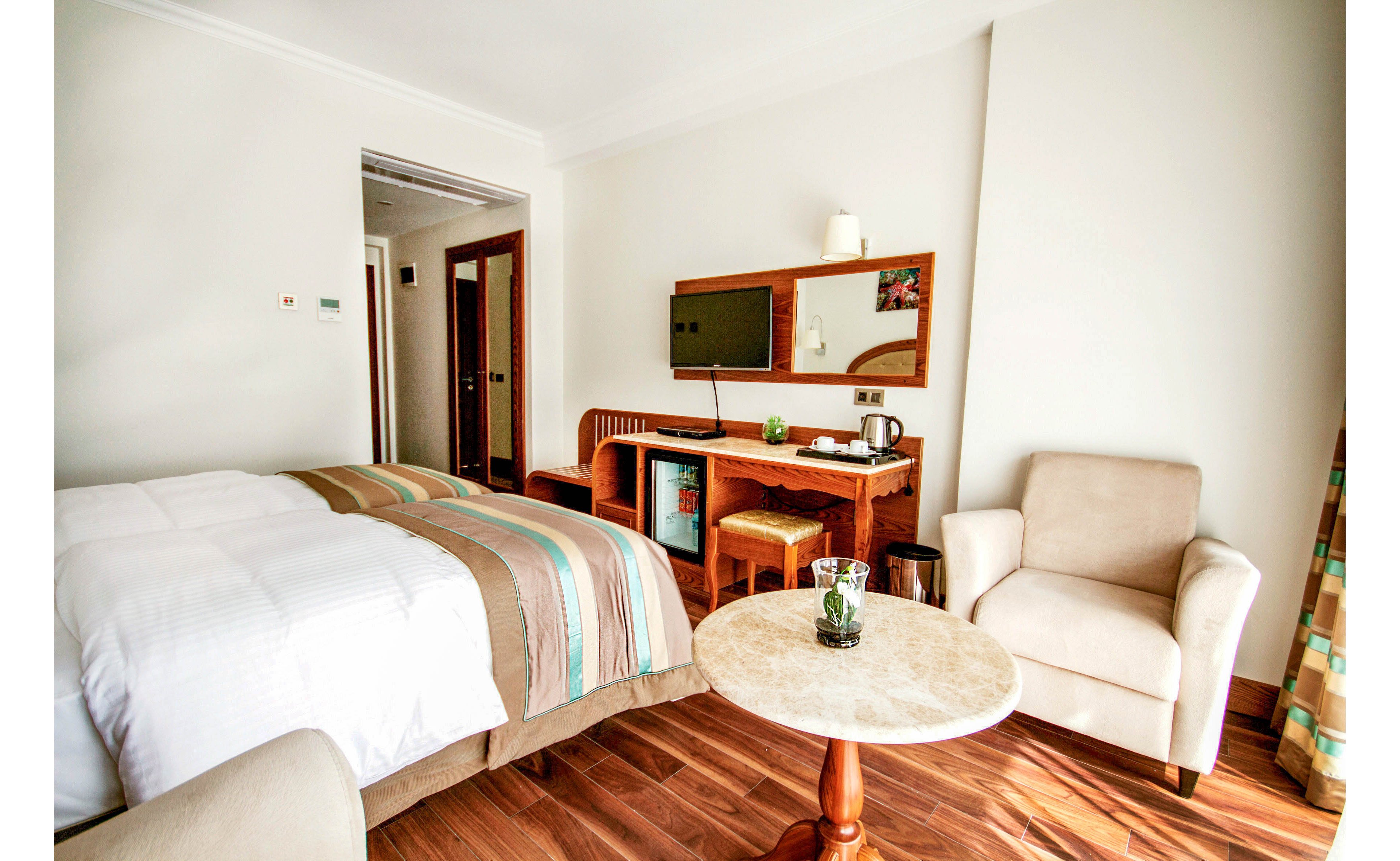 A modern hotel room features a neatly made bed with striped bedding and a small seating area. A round marble-top table with a glass of water and plant sits between a beige armchair and the bed. Along the wall, a wooden desk holds a flat-screen television, mirror, kettle, and cups, with a stool tucked underneath. Warm wooden flooring and neutral-colored walls create a clean, inviting atmosphere. The overall scene suggests a comfortable space for rest and light work during travel.