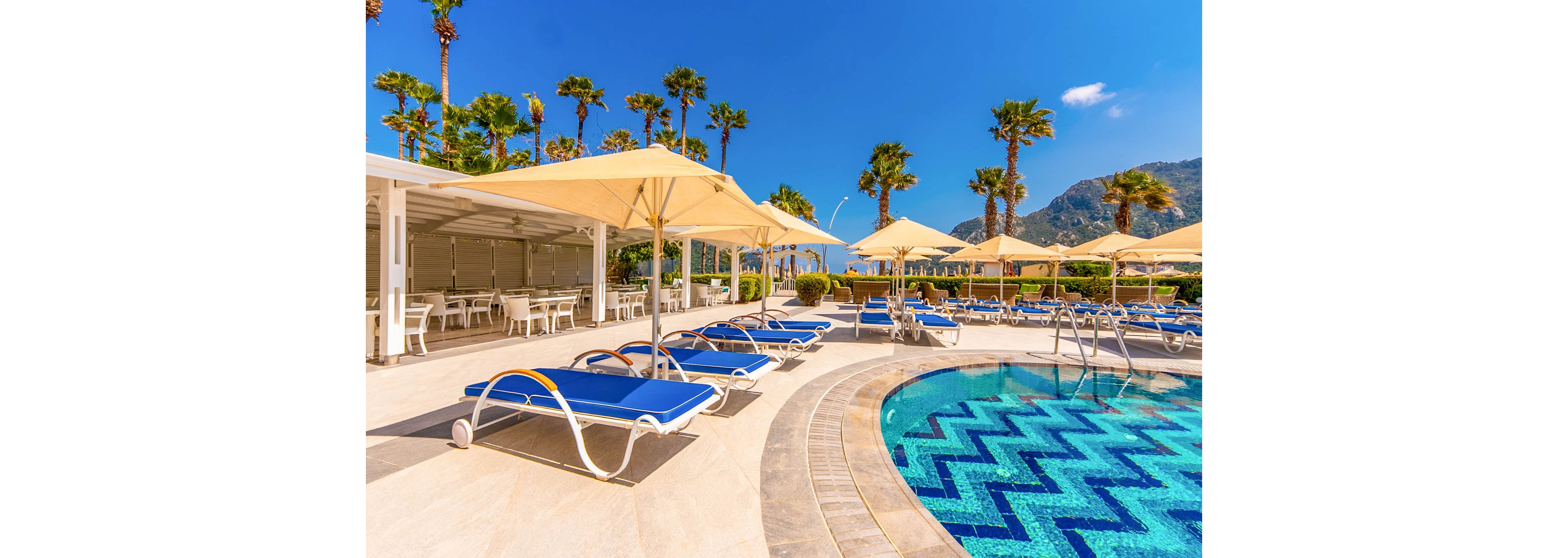 A bright resort pool area features blue lounge chairs arranged neatly under large yellow umbrellas. The swimming pool has a decorative zigzag tile pattern in shades of blue. Palm trees line the background, and a covered outdoor dining area with white tables and chairs sits beside the pool. Clear blue skies and distant mountains create a relaxing vacation atmosphere.