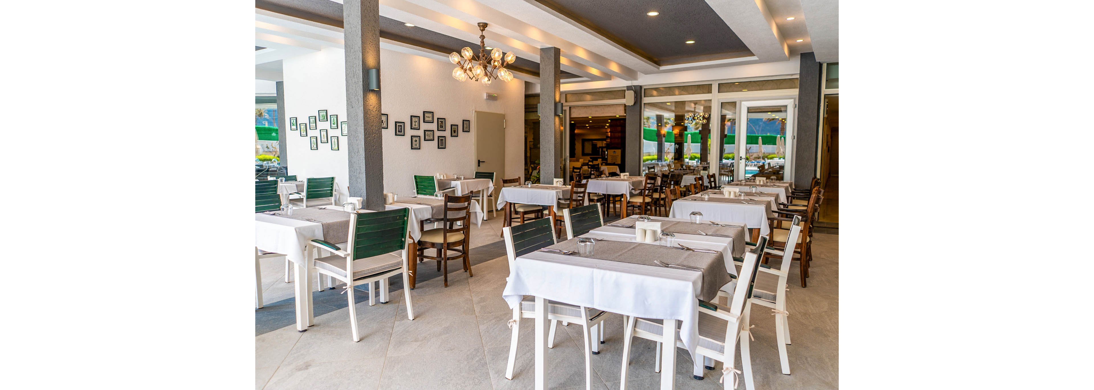 A contemporary restaurant dining area features neatly arranged tables covered with white and gray tablecloths. The space is empty of guests, highlighting rows of chairs and clean place settings. Large windows and glass doors open to an outdoor view, allowing natural light to brighten the interior. Warm ceiling lights and framed wall art add a cozy yet modern ambiance to the room.