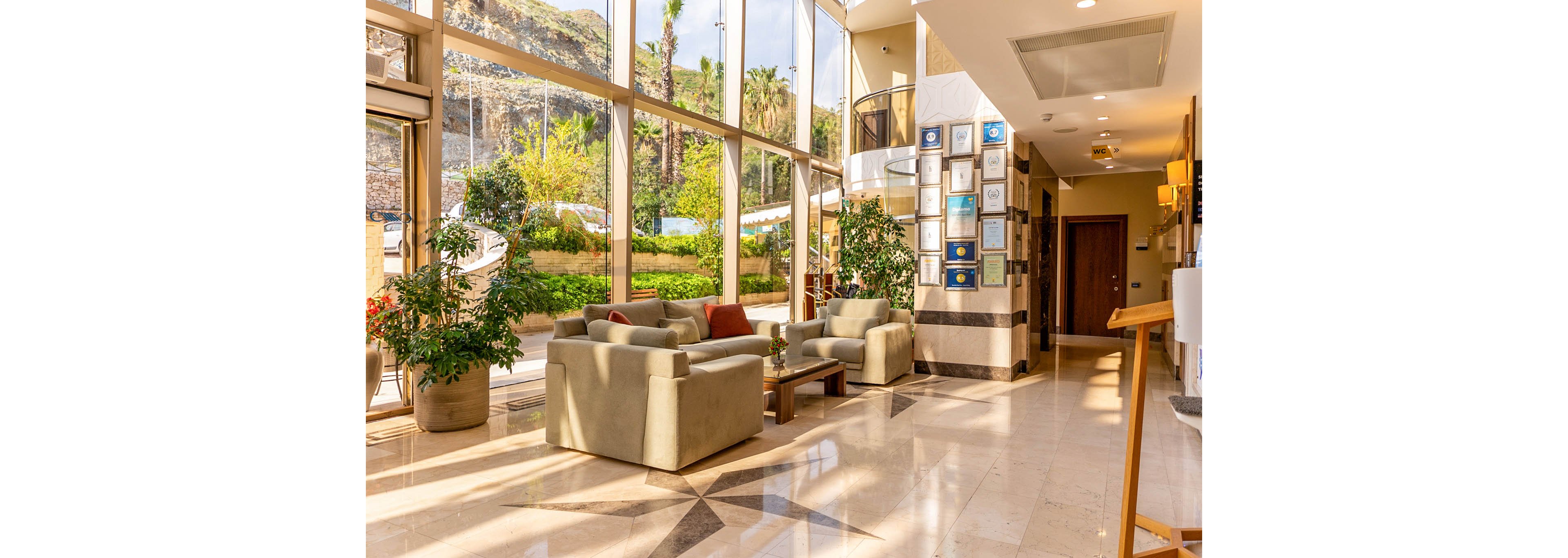 Bright sunlight fills a modern hotel lobby with large floor-to-ceiling windows. Two cushioned armchairs and a sofa are arranged around a low wooden coffee table, creating a comfortable seating area. Potted plants and greenery soften the space and connect the interior to the outdoor landscape visible through the glass. A wall of framed certificates and awards near the corridor adds a professional, upscale touch to the environment.