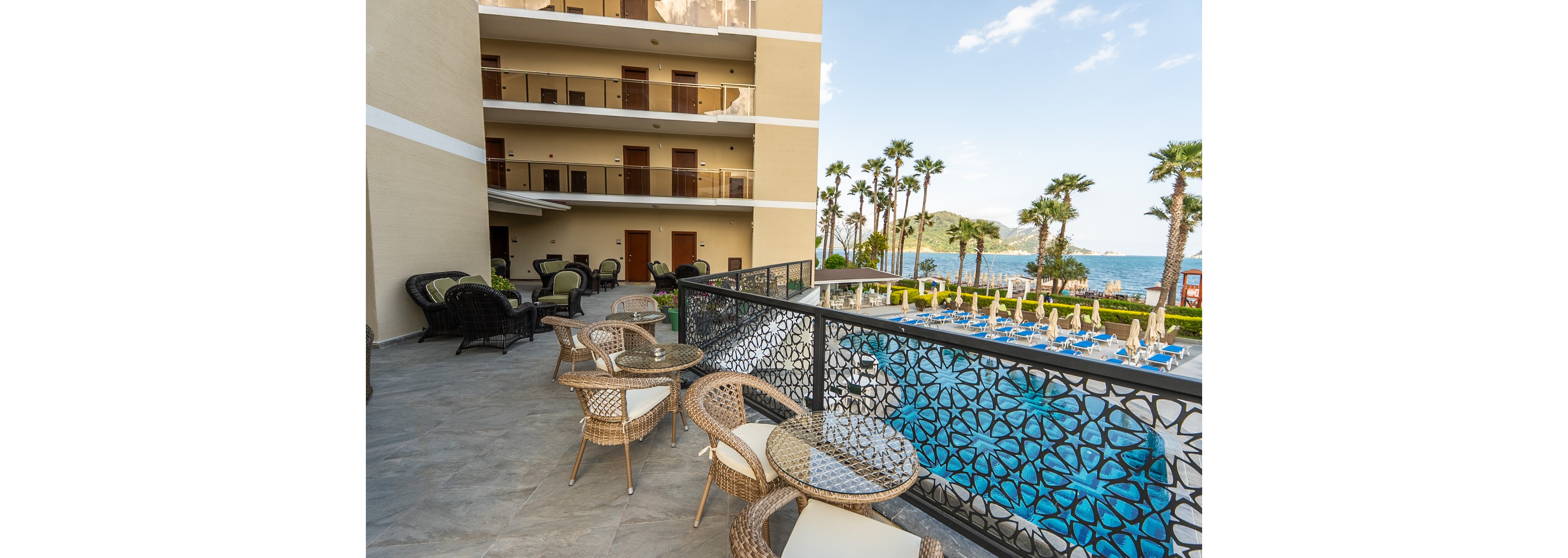 An outdoor hotel balcony features wicker chairs and tables arranged along a decorative metal railing. Below, a large swimming pool with sun loungers and umbrellas stretches toward a line of palm trees. In the background, the blue sea and distant hills create a scenic coastal view. The multi-story hotel building with room balconies frames one side of the scene under a bright, clear sky.