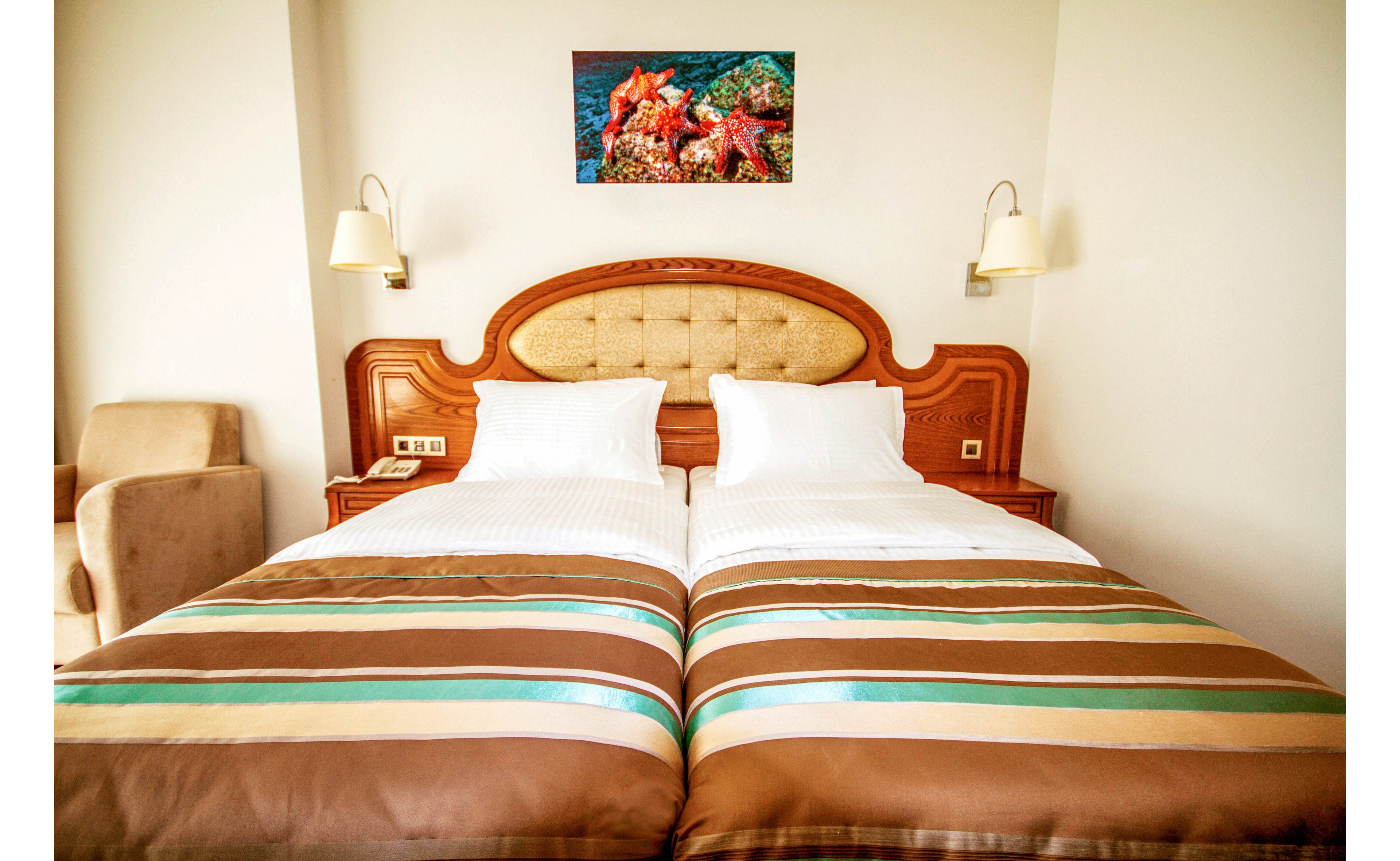 A neatly arranged hotel room features two twin beds pushed together with a classic upholstered wooden headboard. The beds are dressed in white linens and striped brown, teal, and beige bedspreads. Wall-mounted reading lamps flank the headboard, and a framed underwater photograph hangs above it. A beige armchair sits in the corner, contributing to the warm and inviting atmosphere.
