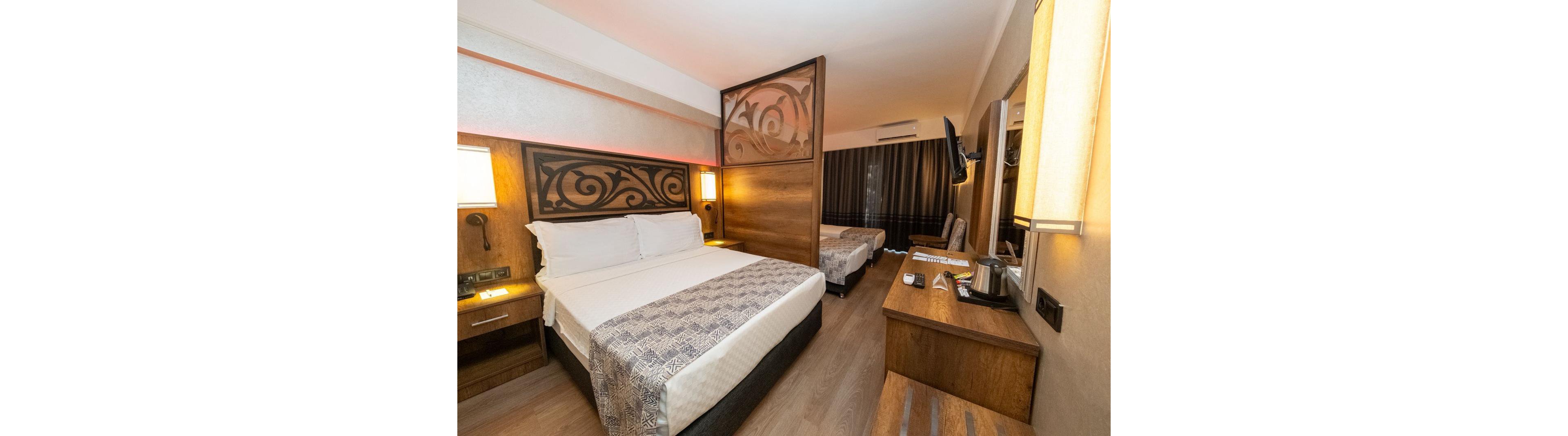 A contemporary hotel room features a large double bed in the foreground with decorative wooden headboard and patterned runner. Another bed is visible behind a partial wooden partition, creating a semi-private sleeping area. The room includes bedside lamps, a desk with phone and kettle, and a wall-mounted television. Warm lighting, wood furniture, and neutral tones create a cozy, inviting atmosphere.