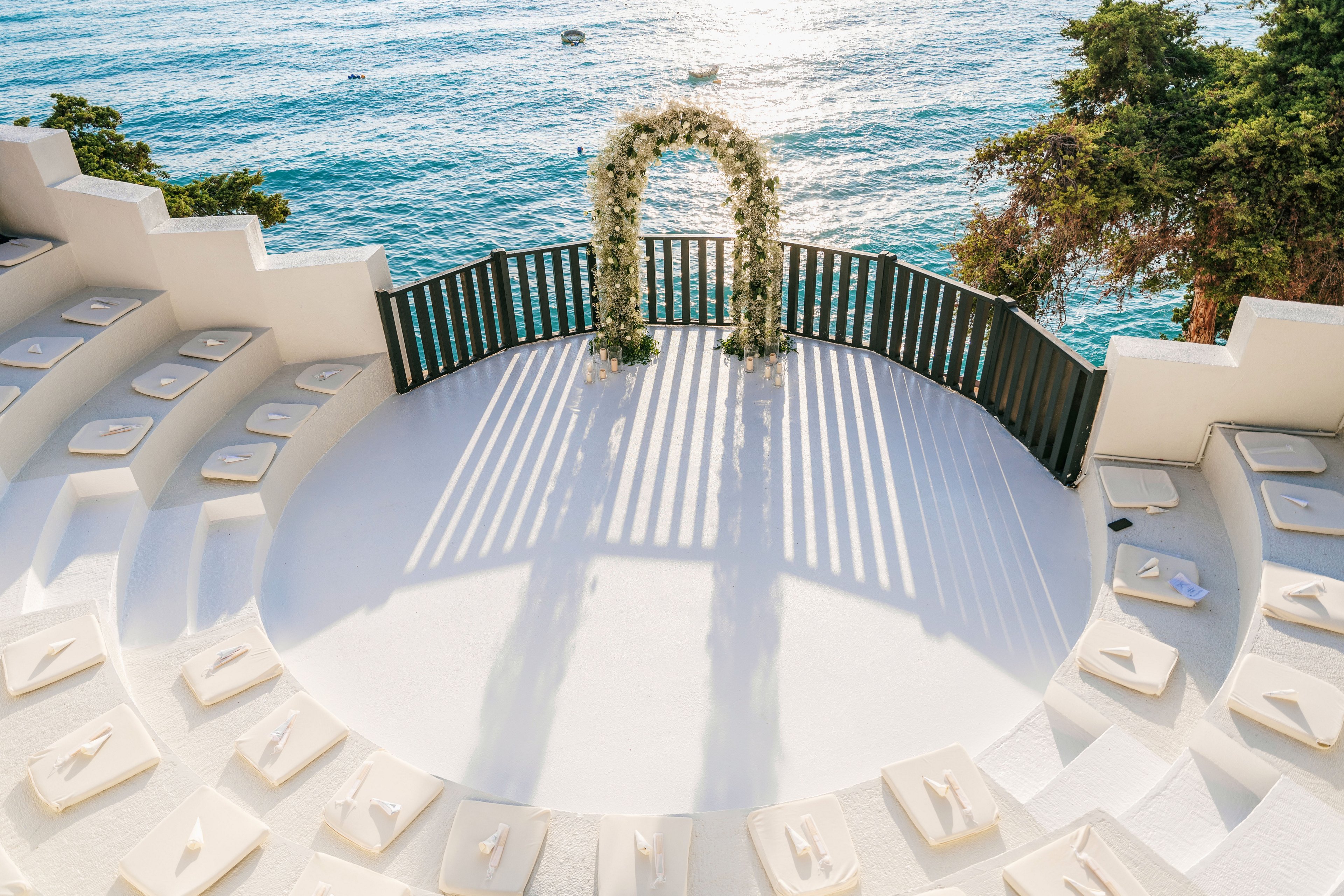 A circular white amphitheater overlooks a calm blue sea, prepared for a wedding ceremony. Cushioned seats are arranged in tiers around the central platform, each with neatly folded napkins. At the edge of the terrace, a floral arch stands in front of a dark railing, casting long shadows on the bright floor. Trees frame the right side of the scene, adding greenery to the coastal setting.