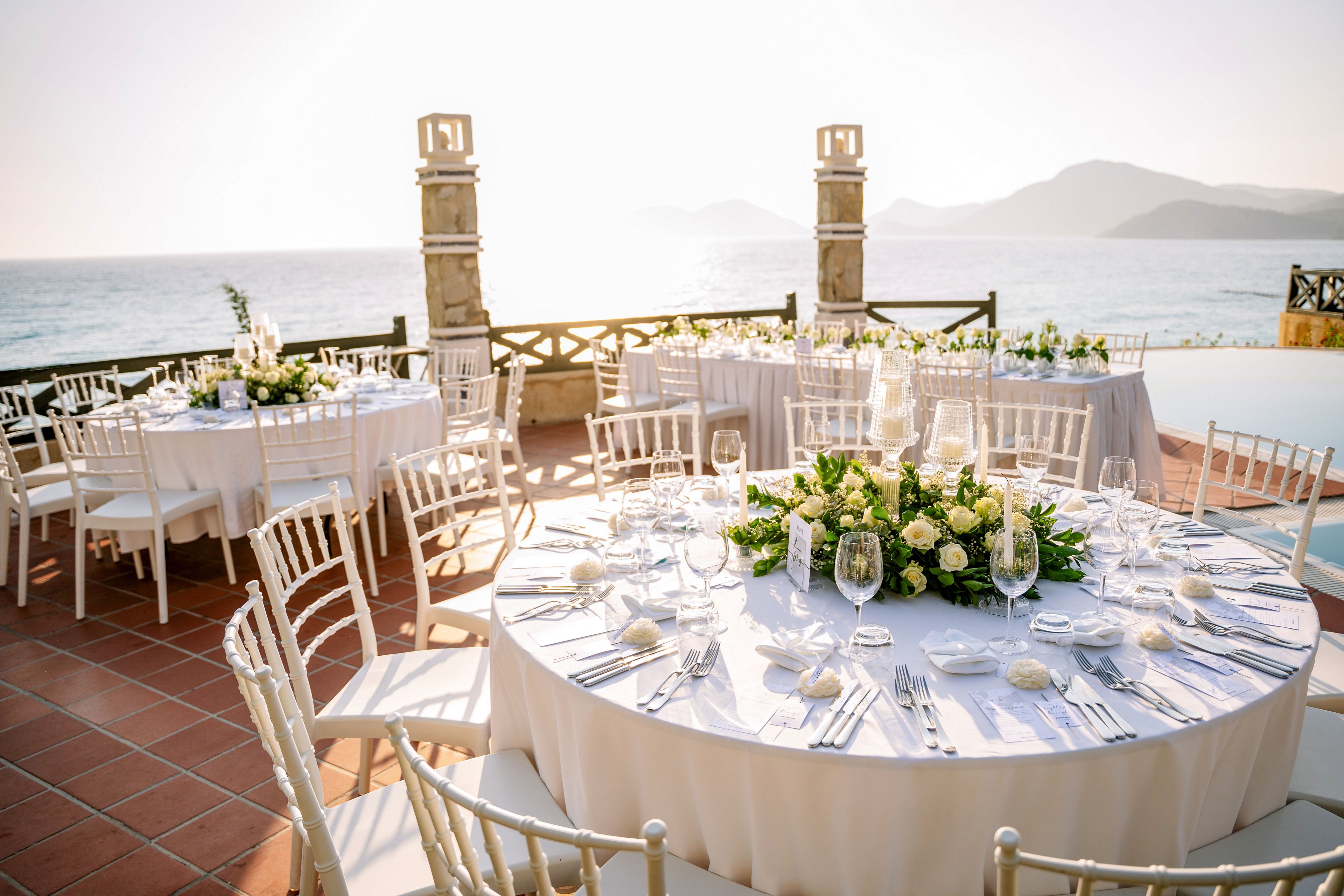 Round banquet tables are set for an elegant outdoor event overlooking the sea. White chairs, crisp linens, and neatly arranged cutlery surround floral centerpieces of white and green blooms. Sunlight reflects off wine glasses and tableware, creating a warm, romantic glow. The terrace setting features a coastal backdrop with calm water and distant mountains, suggesting a wedding or formal celebration.