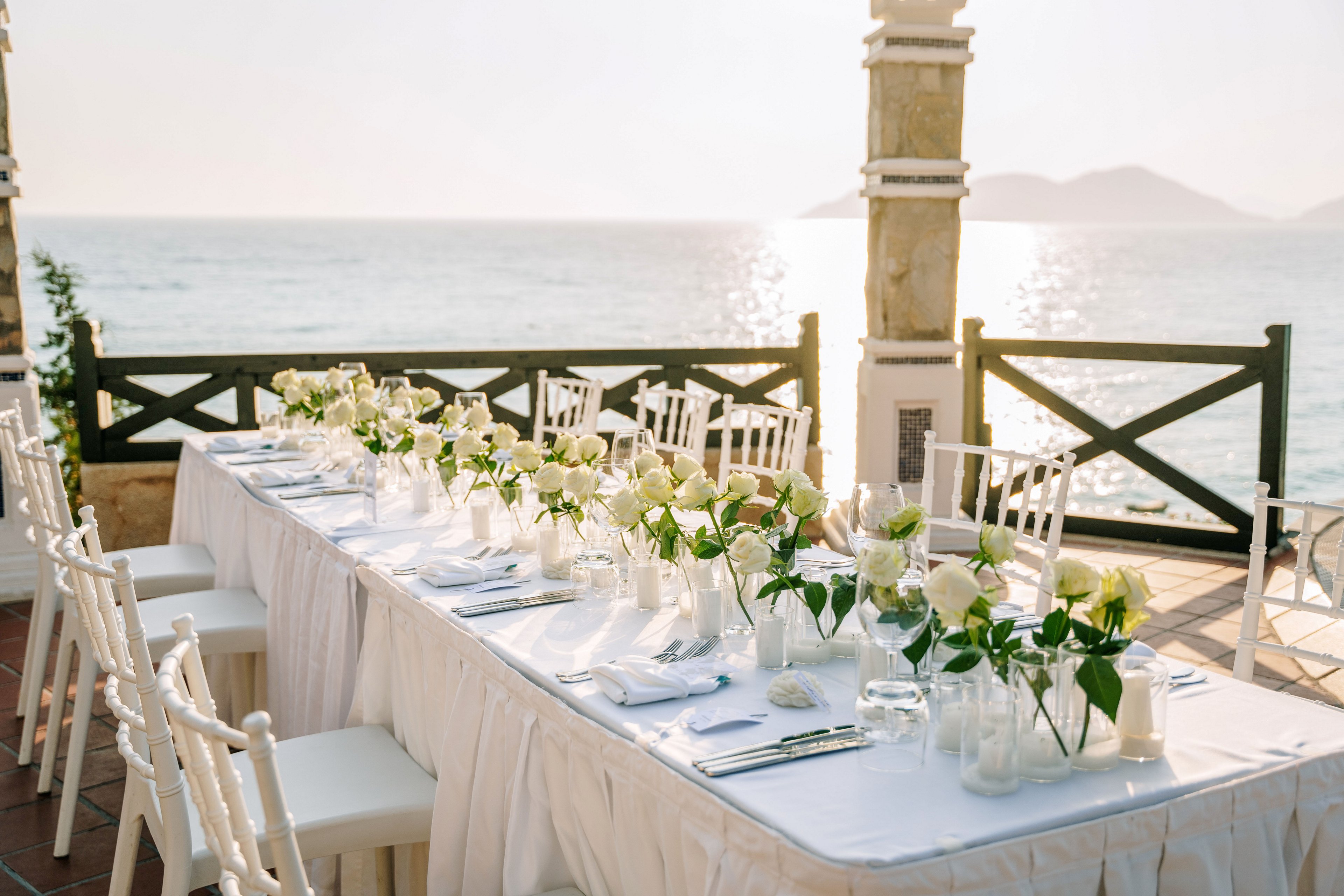 A long, white-clothed table is beautifully arranged for a formal event by the sea. White chairs line both sides of the table, which is decorated with white roses and greenery in glass vases. Glassware, plates, and neatly folded napkins are set for guests, suggesting a wedding or special celebration. The scene is framed by a wooden railing and stone columns, with calm ocean water and distant islands visible in the background. Warm sunlight creates a soft, romantic atmosphere over the entire setting.