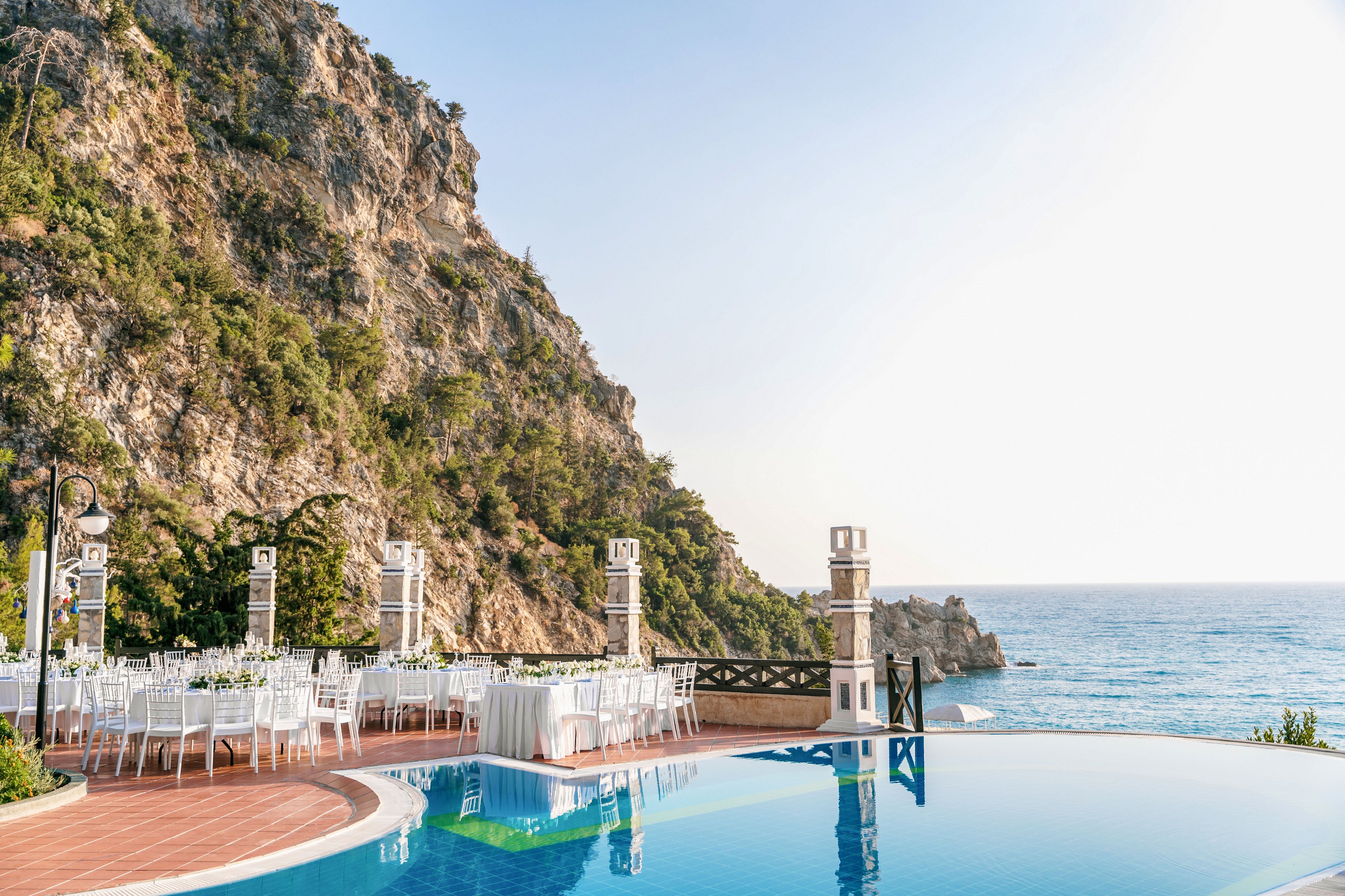 An elegant outdoor wedding reception is set up beside a curved infinity pool overlooking the sea. White chairs and tables are arranged neatly on a tiled terrace, framed by decorative columns and lanterns. A steep rocky cliff covered in greenery rises on one side, while the calm blue ocean stretches to the horizon on the other. The bright daylight and clear sky create a serene, romantic atmosphere.