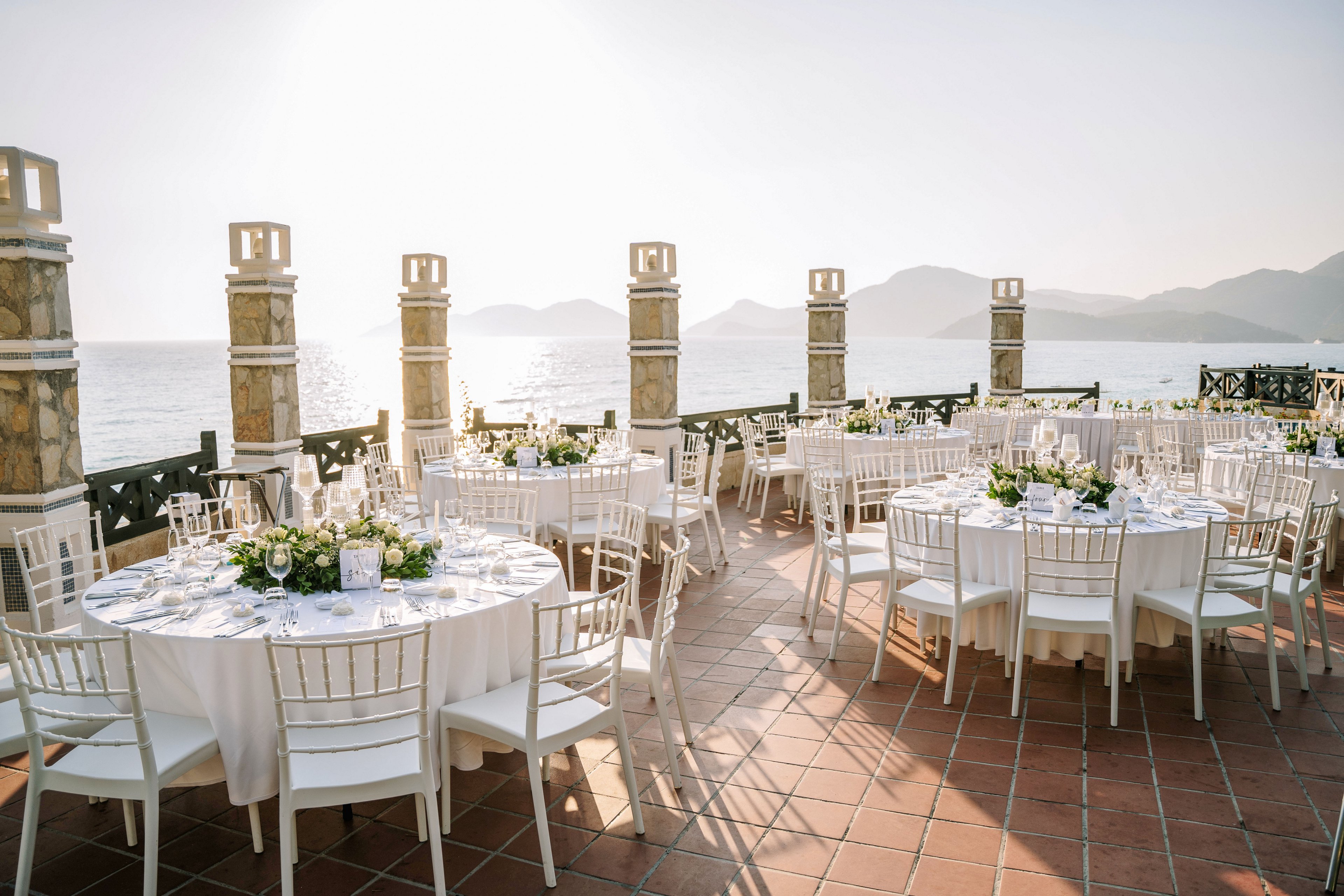 Round tables with white linens and floral centerpieces are arranged on a tiled terrace overlooking the sea. White chiavari chairs surround each table, creating a refined and cohesive event setup. Stone pillars and a decorative railing frame the view of calm water and distant mountains. The soft, warm light suggests a late afternoon or early evening celebration, adding a romantic atmosphere to the outdoor reception.