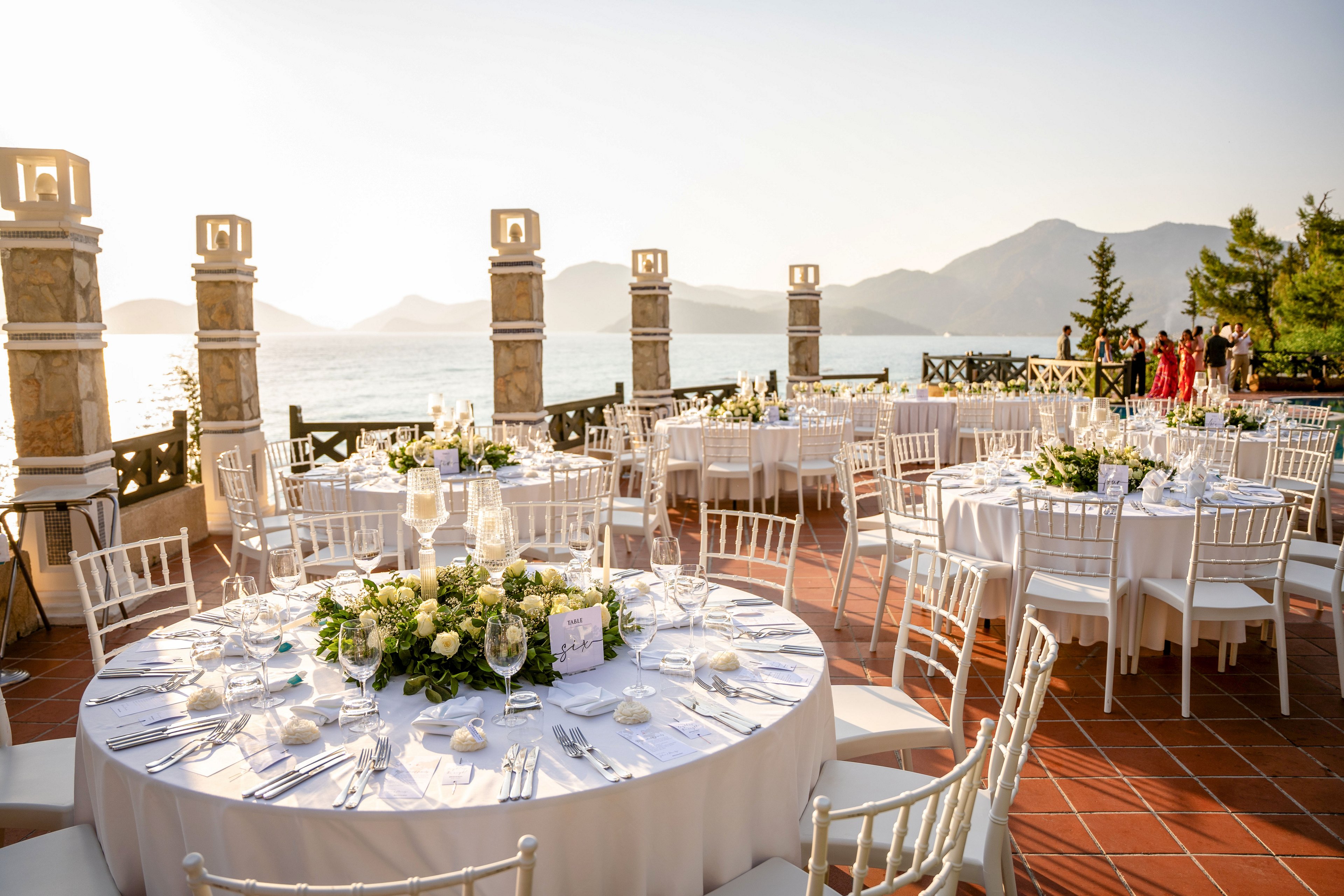 Round tables with white linens and floral centerpieces are arranged on a terrace overlooking a calm sea. White chairs surround each table, set with plates, glasses, and folded napkins for a formal event. Stone columns and railings frame the scenic coastal view with mountains in the distance. Warm sunlight creates a golden, romantic atmosphere as a few guests gather near the edge of the terrace.
