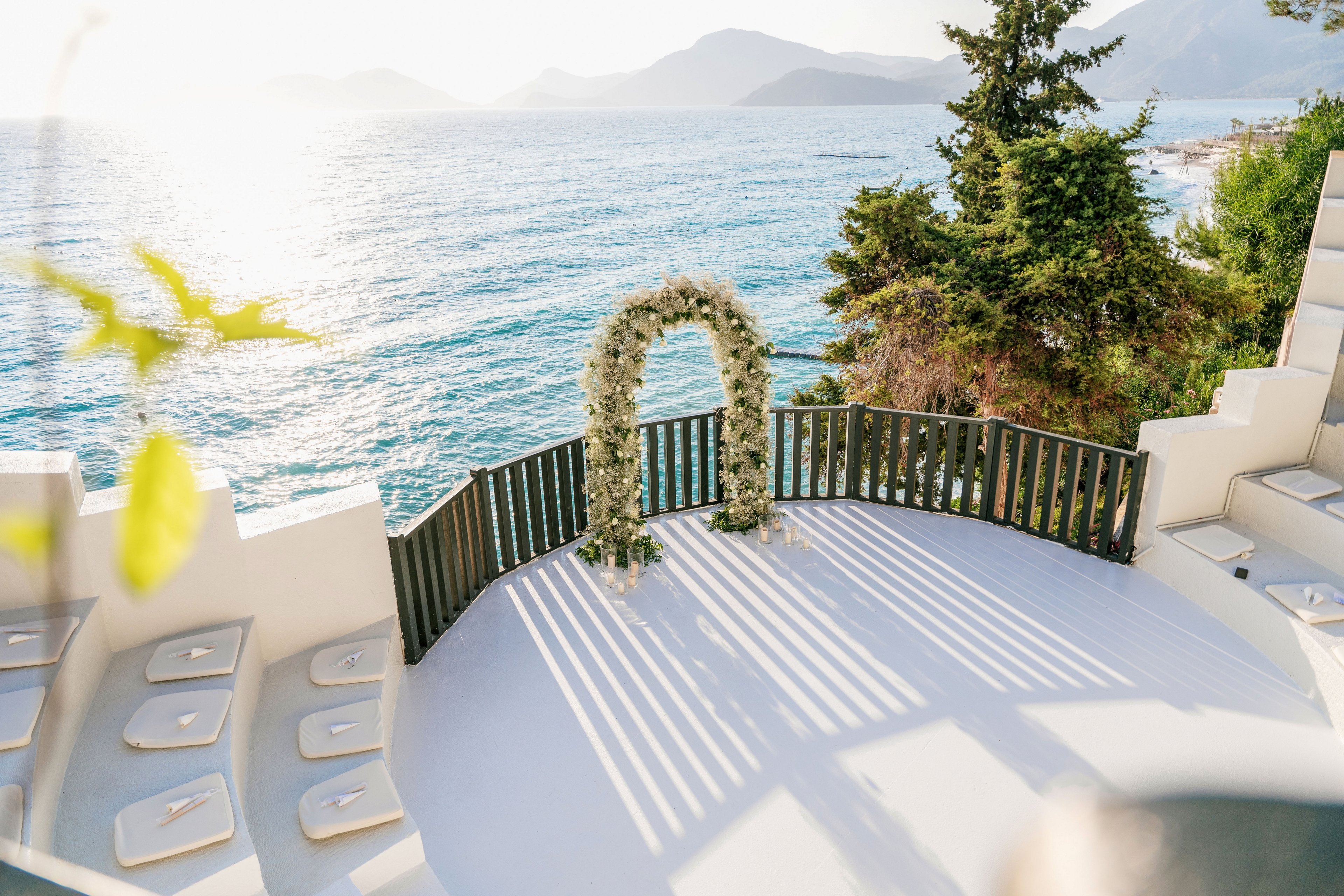 An intimate outdoor wedding ceremony space overlooks a calm, sunlit sea. A floral arch stands at the edge of a white terrace framed by a dark wooden railing. Cushioned seating is arranged neatly along the curved platform, ready for guests. Warm sunlight casts long shadows across the terrace, creating a romantic and serene coastal atmosphere.