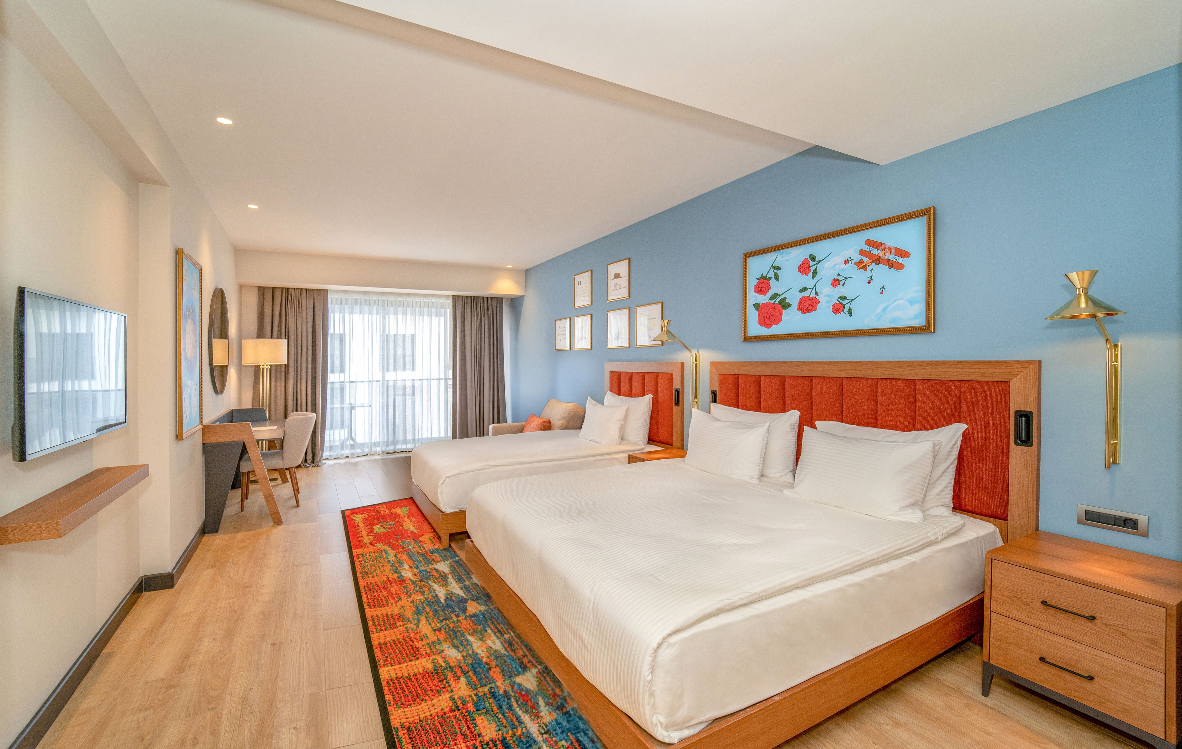 A contemporary hotel room features two large beds with orange upholstered headboards against a light blue accent wall. Colorful framed artwork hangs above the beds, complemented by a patterned runner rug along the wooden floor. A desk and chair sit near the window, which is covered with sheer curtains allowing natural light to fill the space. Warm wood furniture and soft neutral tones create a welcoming, comfortable atmosphere.