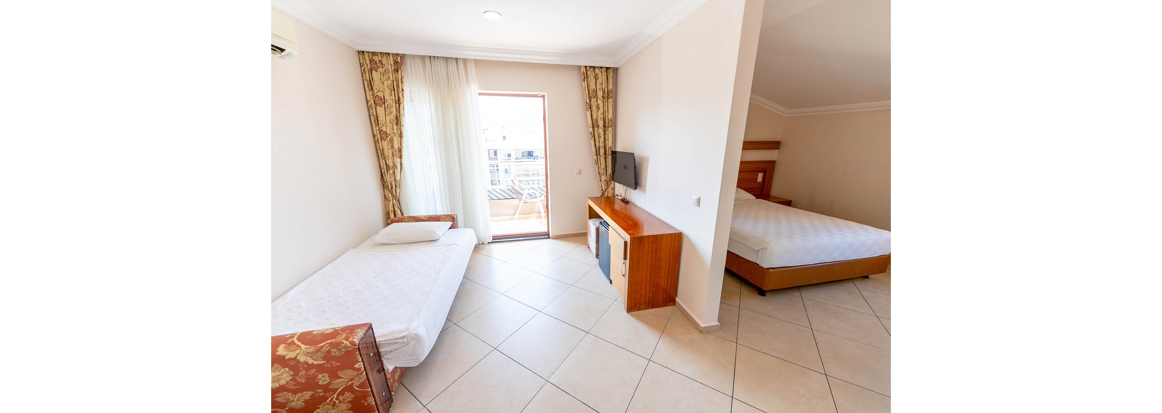 A clean, sunlit hotel room features a single bed in the foreground and a double bed in a partially separated sleeping area. Light tiled flooring and white walls create an airy, spacious feel. A small wooden cabinet holds a flat-screen TV and a compact refrigerator, facing the beds. Sheer curtains and patterned drapes frame a glass door that opens to a balcony with outdoor seating.