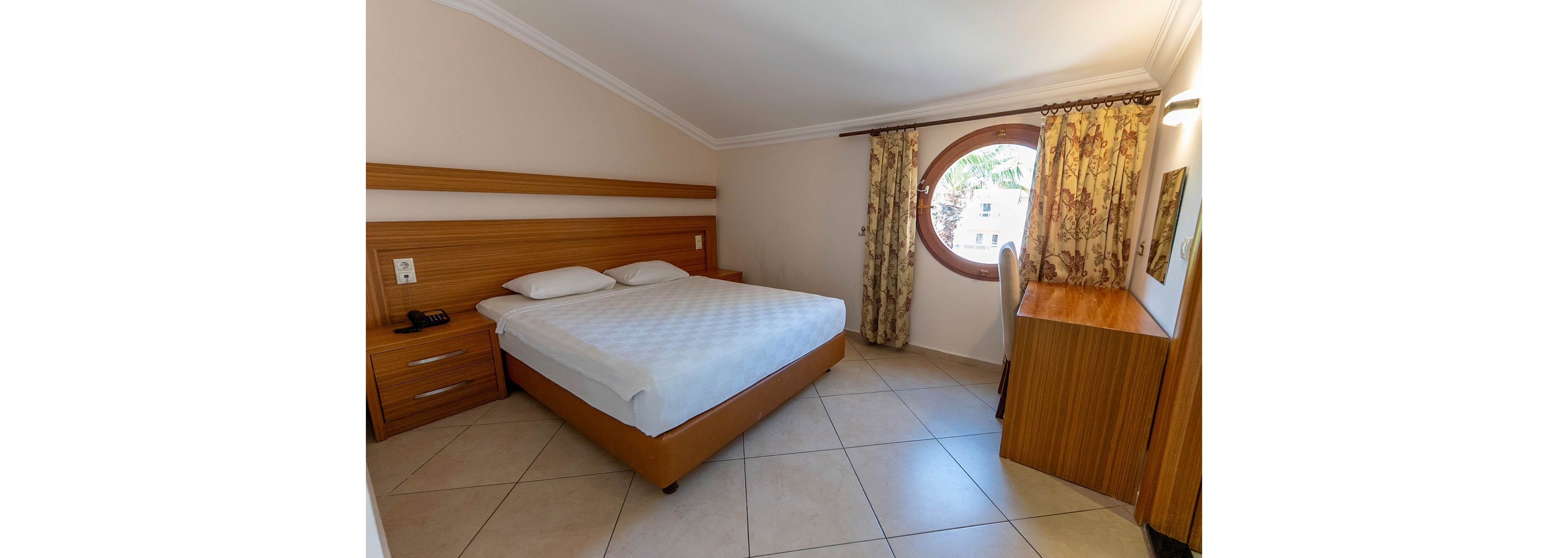 A neatly made double bed dominates a compact hotel bedroom with light tiled flooring. Wooden furniture, including a bedside table and desk, adds warmth to the neutral walls. A large round window with patterned curtains lets in natural light and offers a view of greenery outside. The room appears clean, simple, and functional, suitable for travelers seeking a comfortable stay.