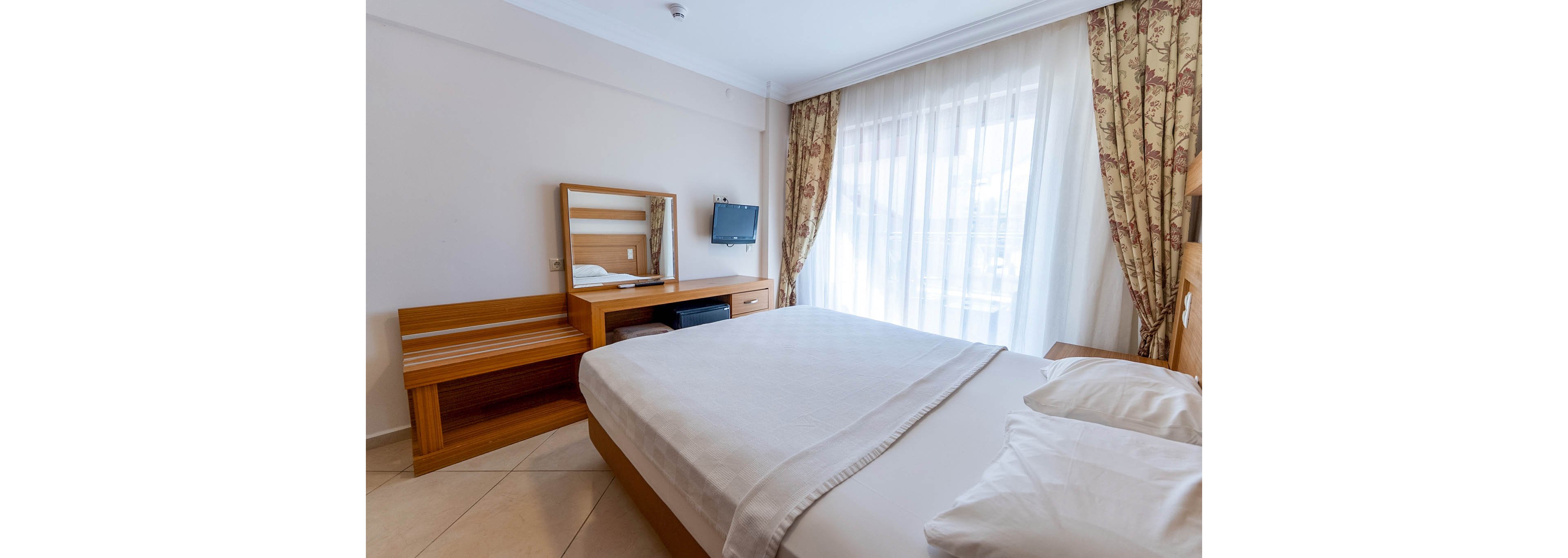 A neatly made double bed dominates a bright hotel bedroom with light-colored walls and tiled flooring. Natural light streams through large sheer curtains covering a window or balcony door, creating an airy atmosphere. A wooden desk with a mirror and bench sits along one wall, accompanied by a small wall-mounted television and a compact cabinet. The overall scene feels clean, simple, and inviting, suitable for guest accommodation.