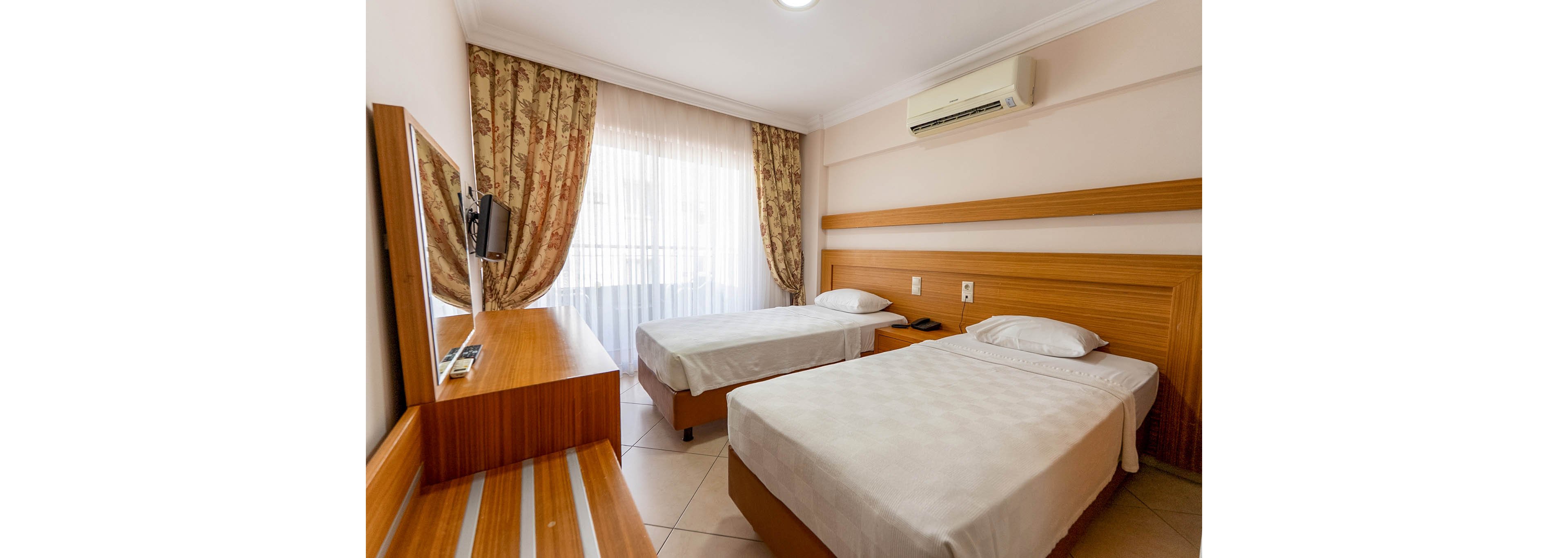 A tidy hotel room features two single beds with white linens against a wooden headboard. Large sheer curtains cover a window, allowing soft daylight to fill the space. A wall-mounted air conditioner is positioned above the beds, and a small flat-screen television faces them from a wooden desk. The room has light-colored tiled flooring and a simple, minimalist decor with neutral tones.