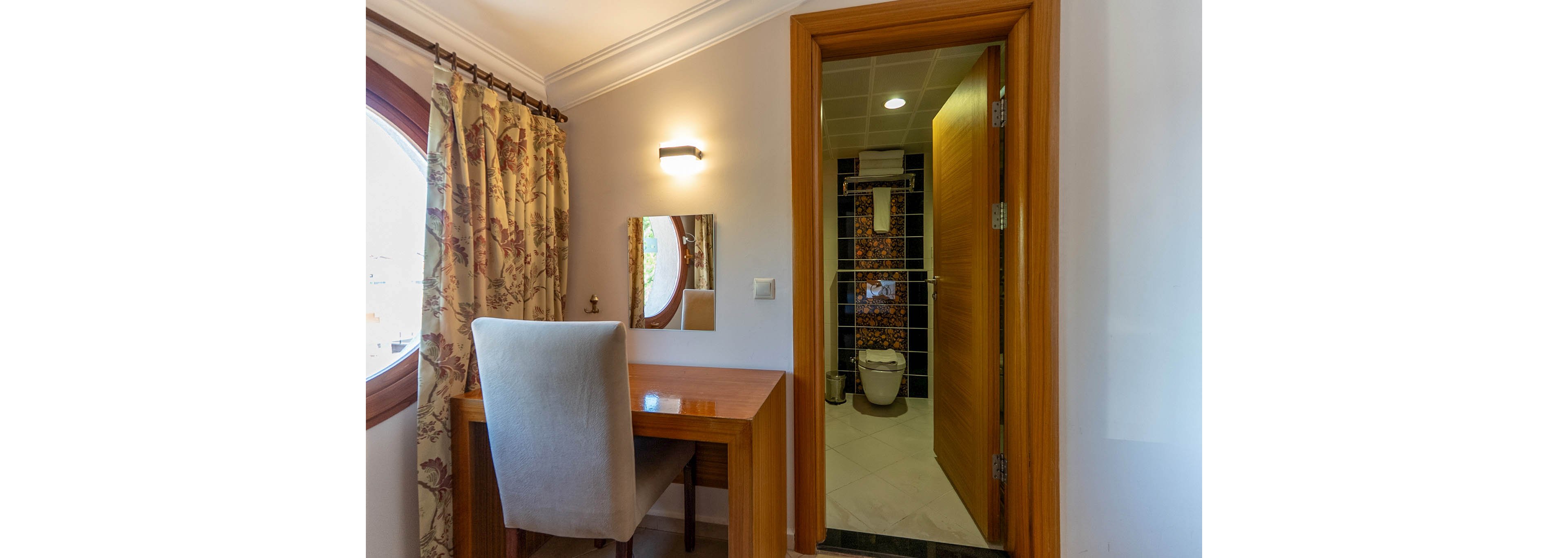 A small hotel room features a wooden desk with a cushioned chair positioned beside a round window with patterned curtains. A wall-mounted light and mirror create a simple workspace or vanity area. The open door reveals a compact bathroom with a toilet, tiled walls, and a towel hanging above. Warm wood tones and soft lighting give the space a comfortable, inviting feel.