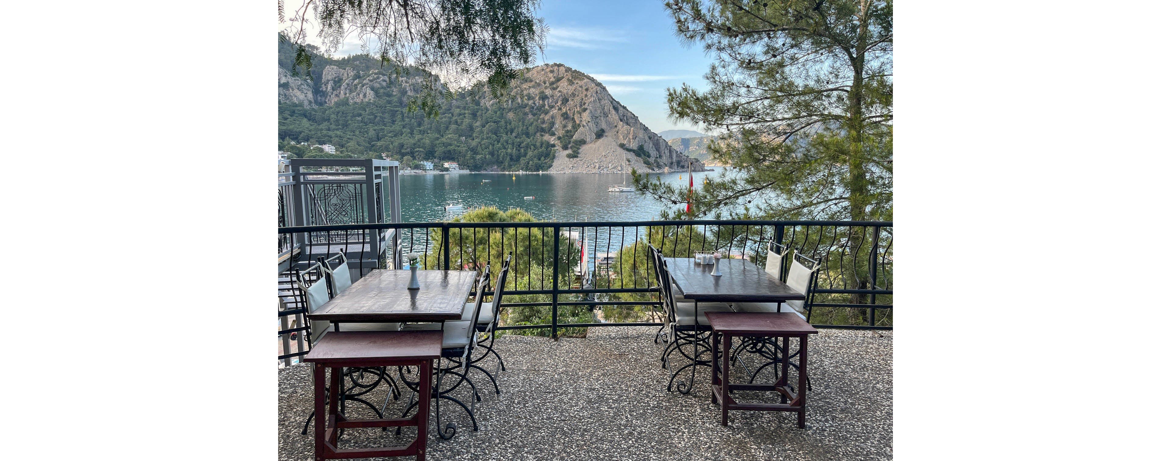 An outdoor café terrace with empty tables and chairs overlooks a calm coastal bay. The seating area is bordered by a black metal railing on a stone or gravel floor. Across the water, a rocky hillside covered with trees rises prominently under a clear blue sky. Boats and small structures are visible along the shoreline, adding detail to the scenic waterfront. Tall trees frame the view, creating a relaxed and inviting atmosphere.