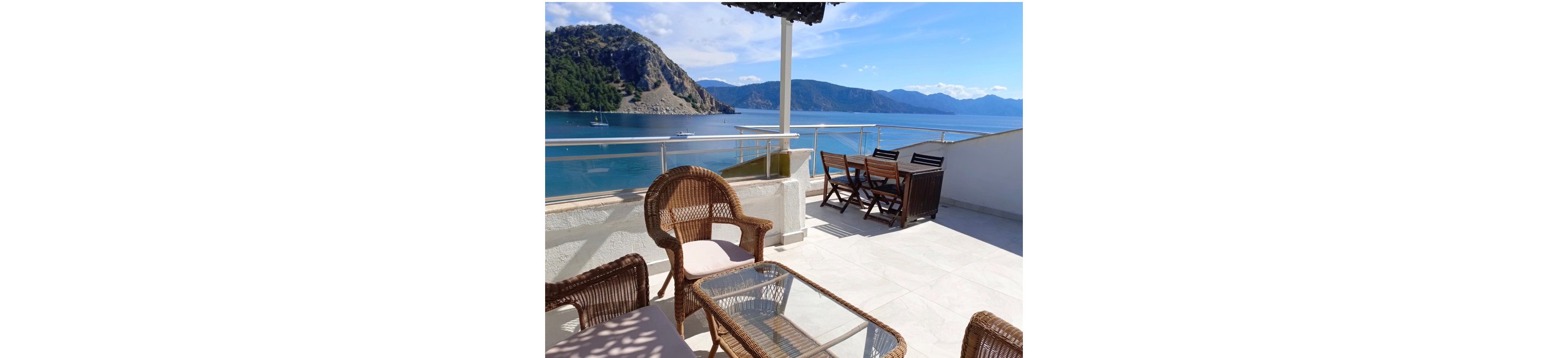 An open terrace overlooks a calm blue sea and distant mountains under a bright sky. Wicker chairs and a glass-top table sit in the foreground, while a wooden dining set stands near the railing. The scene suggests a relaxing coastal retreat with boats anchored in the water. Sunlight highlights the clean tiled floor and the inviting outdoor seating area.
