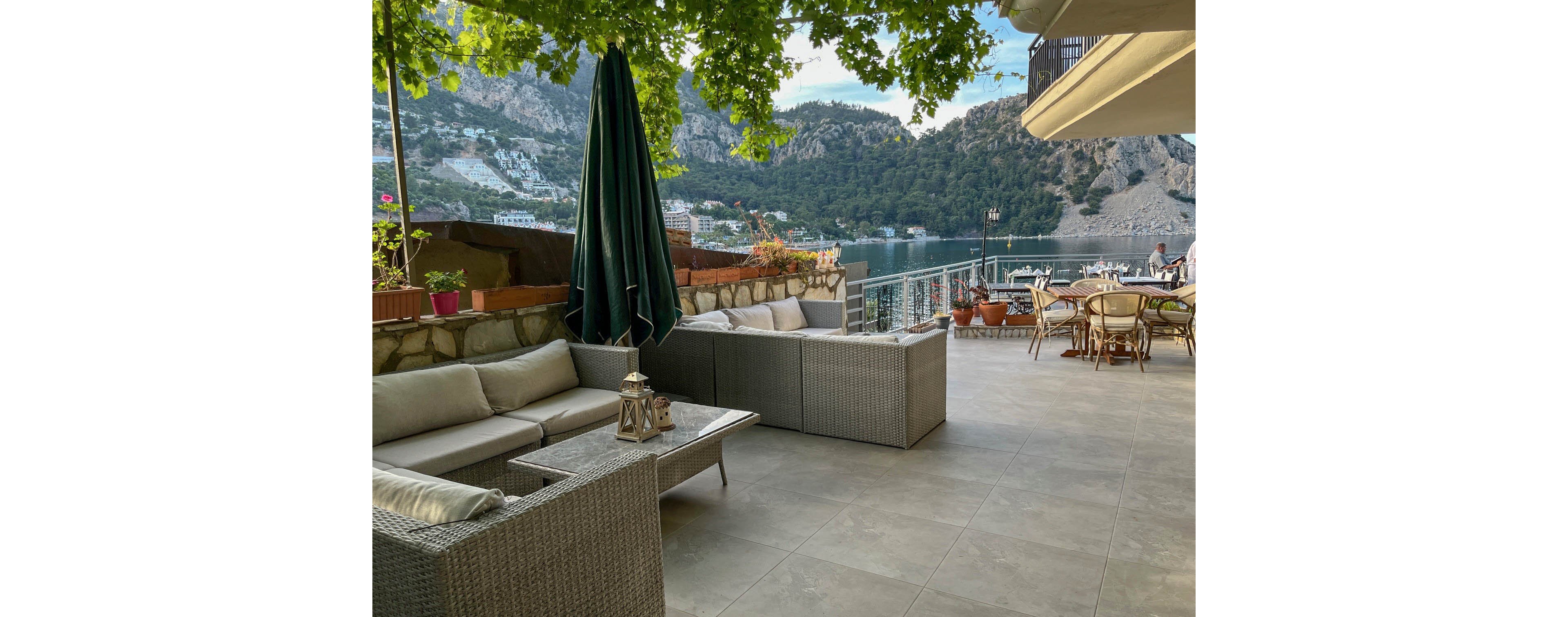An outdoor terrace lounge overlooks a calm lake framed by steep, rocky mountains. Wicker sofas with light cushions and decorative lanterns create a comfortable seating area under leafy green vines. A row of tables and chairs lines the railing, offering views of the water and distant hillside buildings. The scene feels peaceful and inviting, ideal for relaxed dining or quiet conversation.