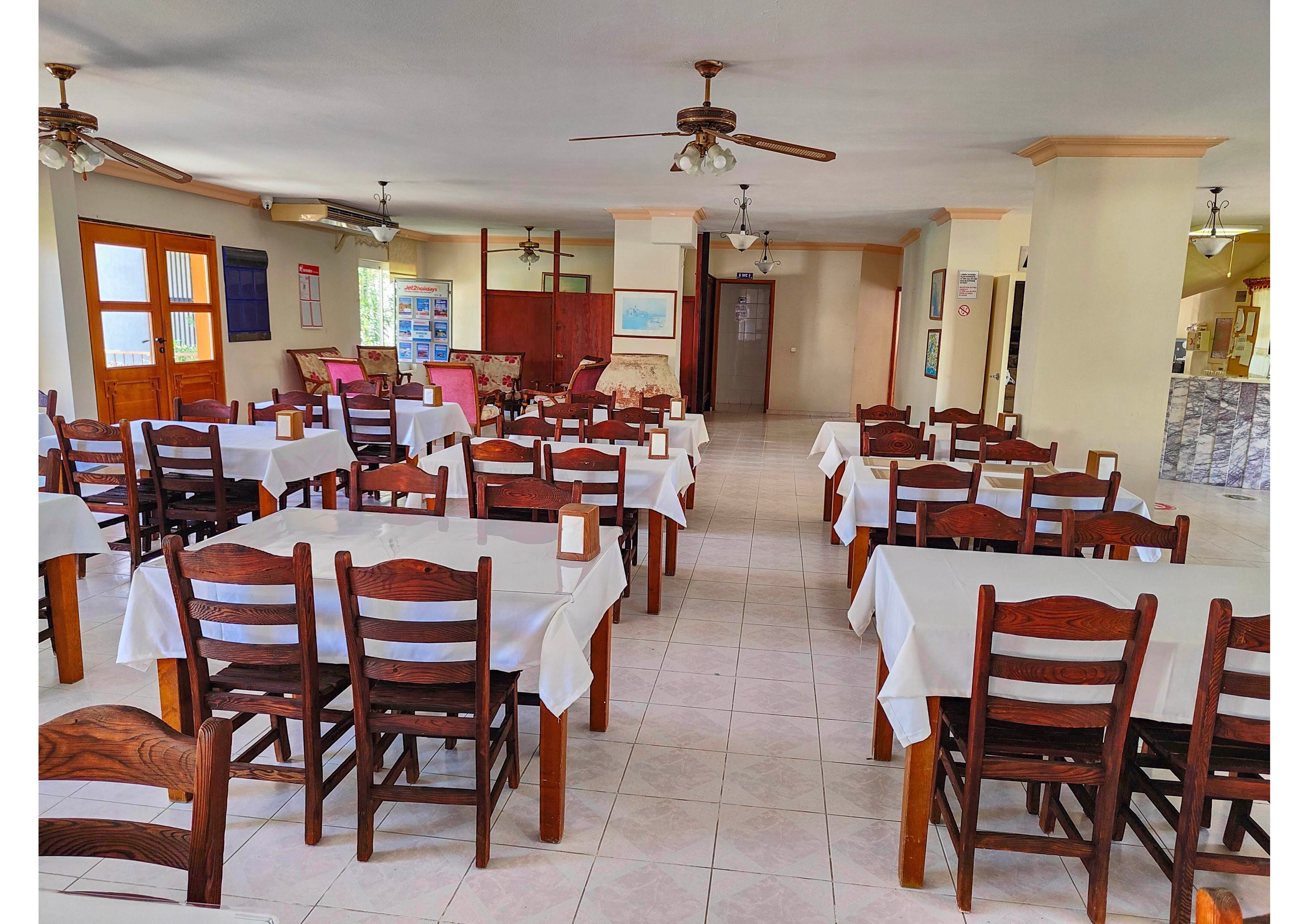 A bright, empty restaurant dining room features rows of wooden chairs and tables covered with white tablecloths. The space is open and airy, with tiled floors and ceiling fans providing ventilation. Natural light enters through large windows and doors on the left side, while the back wall shows additional seating and decor. The overall atmosphere is clean, simple, and ready for guests.