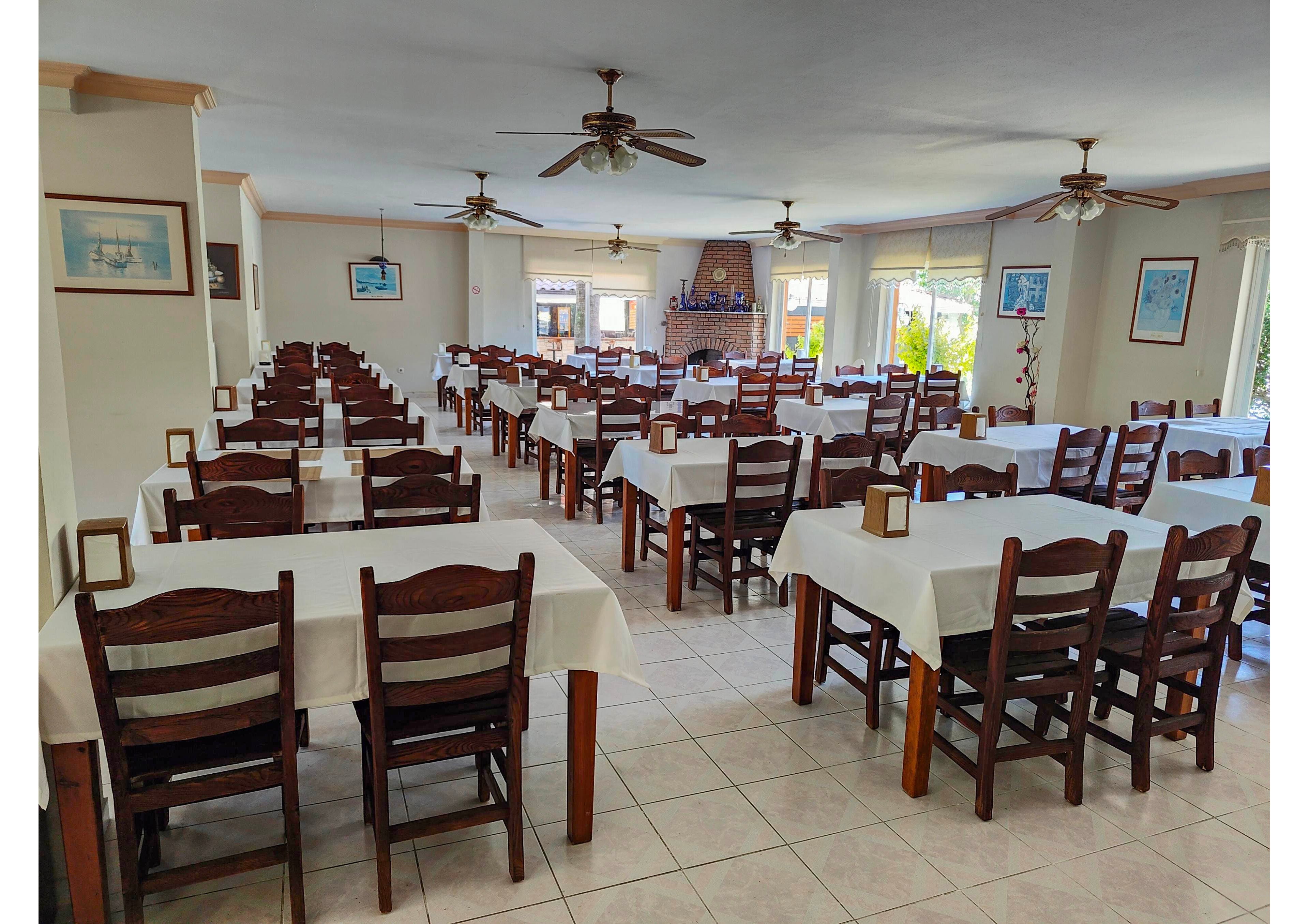 A spacious restaurant dining room is filled with rows of wooden tables and chairs covered in white tablecloths. The room is empty of people, creating a calm and orderly atmosphere. Large windows allow natural light to brighten the tiled floor and white walls, which are decorated with framed artwork. Ceiling fans and simple table lanterns add to the casual, welcoming setting.