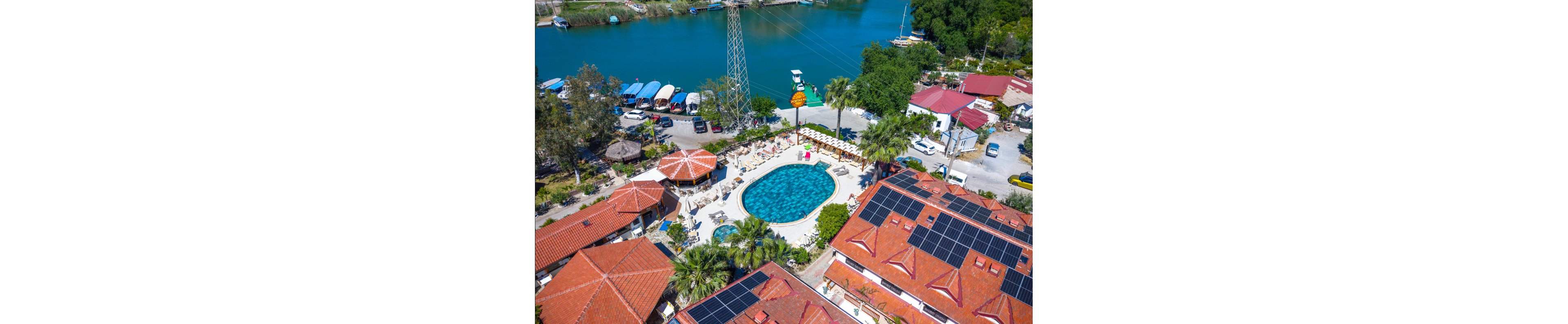 An aerial view shows a riverside resort centered around a large circular swimming pool. Sun loungers and umbrellas surround the pool, with guests relaxing in the sunshine. Red-tiled buildings with rooftop solar panels frame the scene, and lush palm trees add greenery. A calm river with moored boats and a small dock runs alongside the property, creating a tranquil vacation atmosphere.