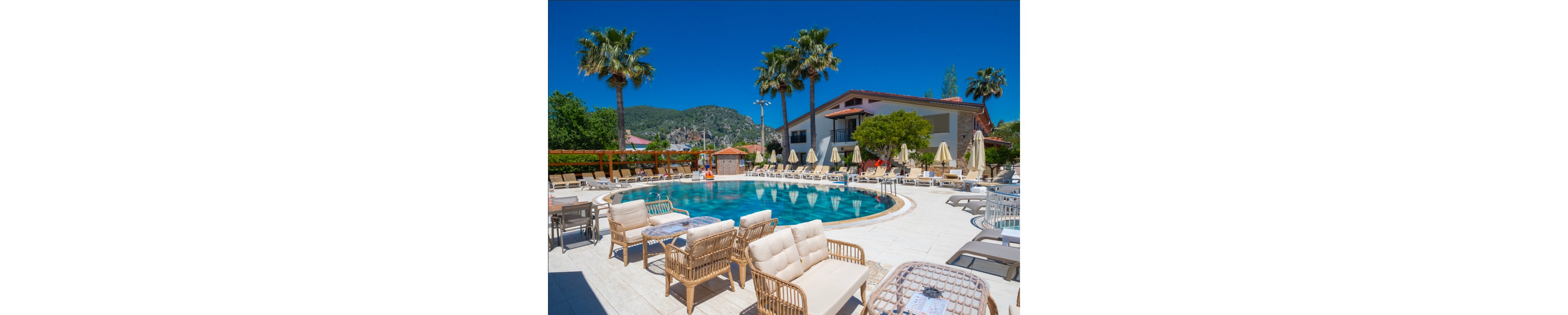 An outdoor resort swimming pool is surrounded by wicker lounge chairs and umbrellas under a clear blue sky. Palm trees and landscaped greenery frame the pool area, creating a tropical atmosphere. A low-rise hotel building with balconies stands in the background against a backdrop of forested hills. The bright sunlight and vivid colors emphasize a relaxed vacation setting.