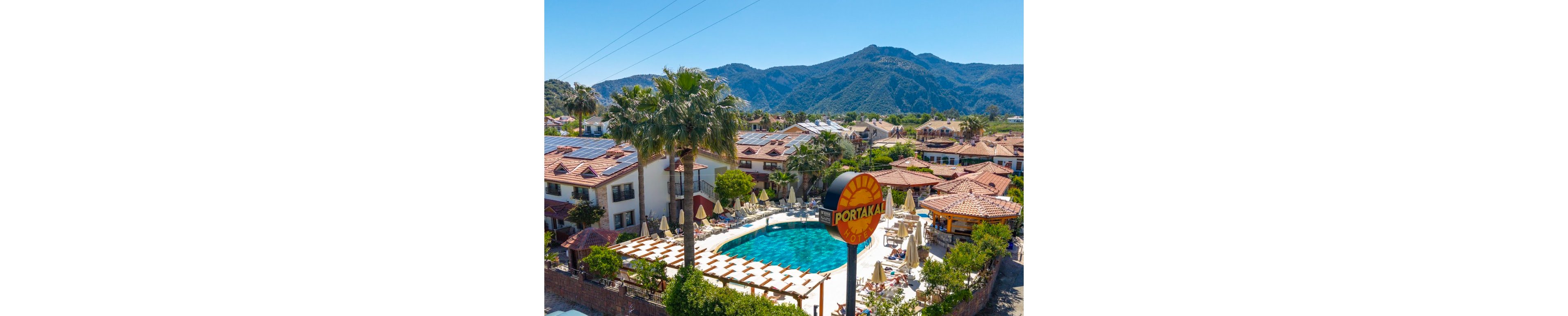 An outdoor hotel pool area is surrounded by sun loungers, umbrellas, and palm trees within a Mediterranean-style resort. The buildings feature terracotta roofs and some have solar panels installed on top. A round sign reading "PORTAKAL" stands prominently near the pool. In the background, lush green mountains rise under a clear blue sky, creating a scenic vacation atmosphere.