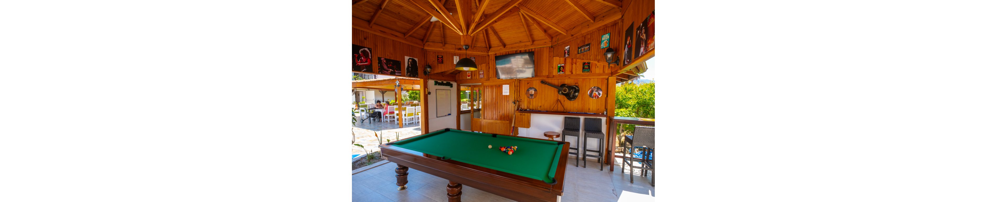 A green-felt billiard table stands in the center of a wooden, open-air bar area. The interior features a wooden ceiling, bar counter with high stools, and a mounted guitar on the wall. Framed music posters and a large television decorate the walls, adding a casual entertainment vibe. In the background, people sit at white tables in an outdoor dining space surrounded by greenery.