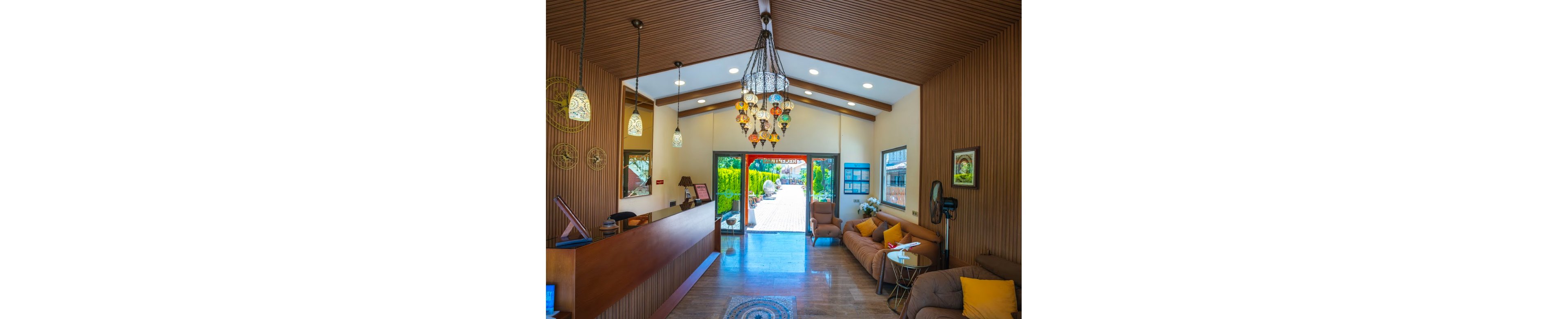 A bright hotel lobby features a wooden reception desk and slatted wood walls leading toward a glass doorway that opens to a sunny courtyard. Colorful hanging lanterns and ceiling lights illuminate the space, creating a warm and inviting atmosphere. Comfortable sofas with yellow cushions line the right side, accompanied by small side tables and decorative accents. Large windows and the open door reveal greenery and outdoor seating, enhancing the welcoming feel of the interior.