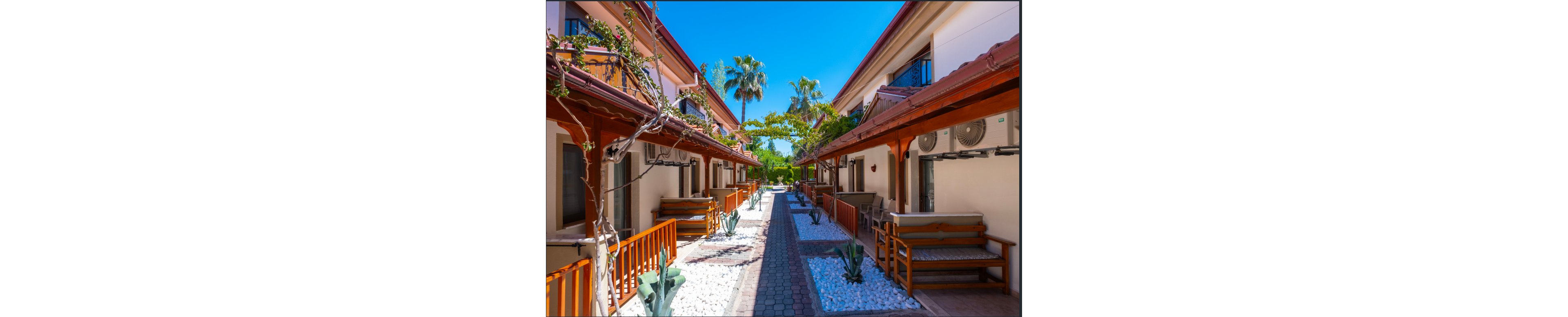 A bright pedestrian walkway runs between two rows of resort-style guest houses. Wooden benches and railings line the fronts of the buildings, with air conditioning units mounted above. Decorative white stones and small desert plants border the paved path, leading the eye toward lush greenery and palm trees in the distance. The clear blue sky and vibrant colors create a warm, inviting vacation atmosphere.