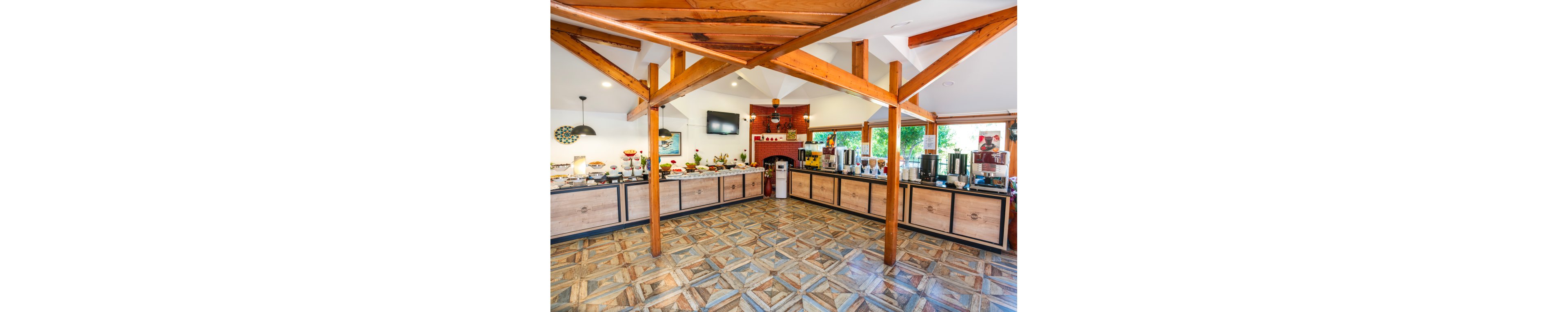 A spacious self-service buffet area is shown with long counters filled with food and beverage dispensers. The room features prominent wooden support beams and patterned tile flooring. Large windows and bright lighting create an airy, welcoming atmosphere. A wall-mounted television and decorative elements add a modern touch to the dining space.