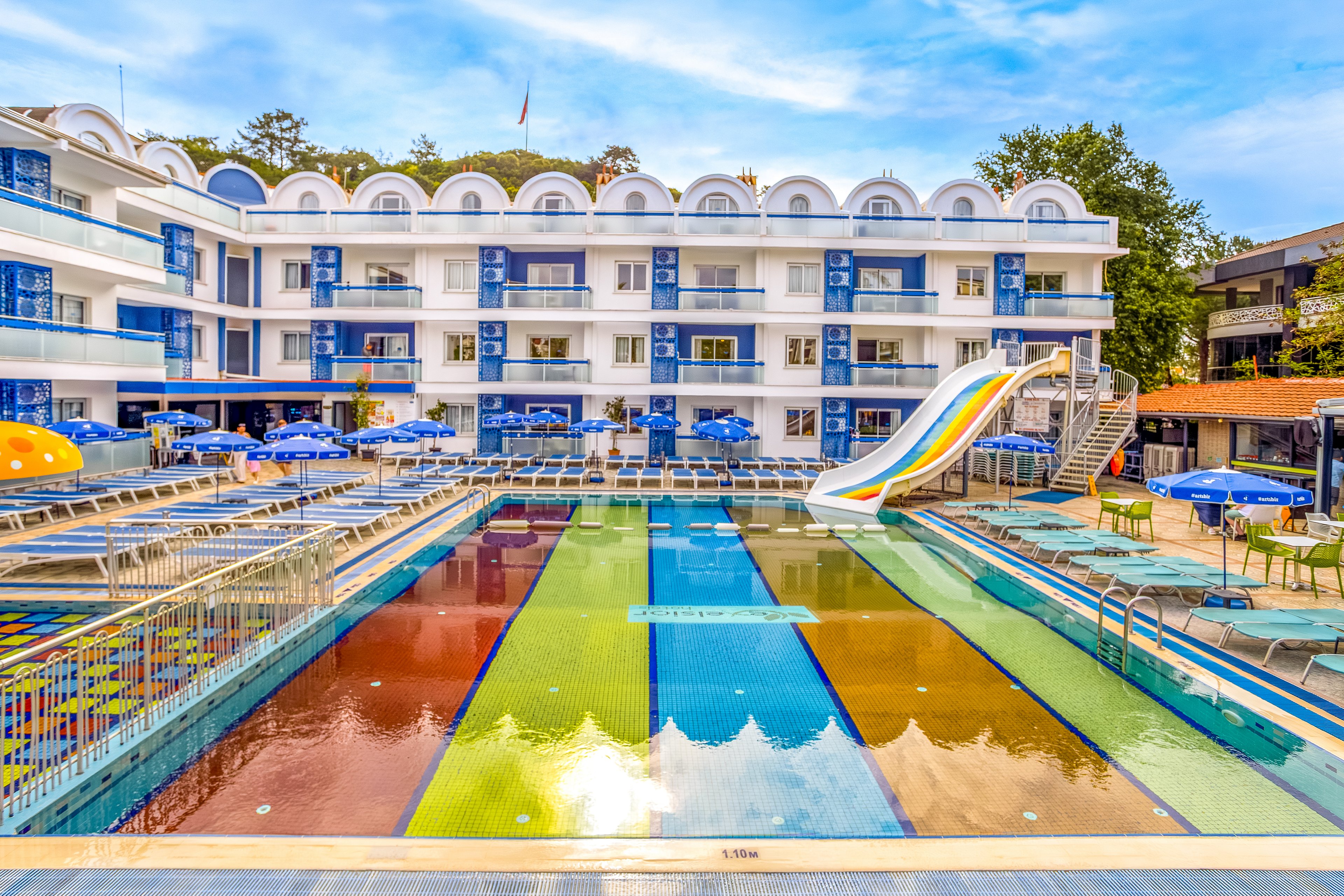 A vibrant outdoor hotel pool area features multicolored pool lanes in front of a modern white and blue building. Numerous sun loungers and blue umbrellas surround the pool, creating a lively resort atmosphere. A curved waterslide descends into the pool, emphasizing family-friendly fun and recreation. The sky is bright and clear, enhancing the cheerful holiday setting.