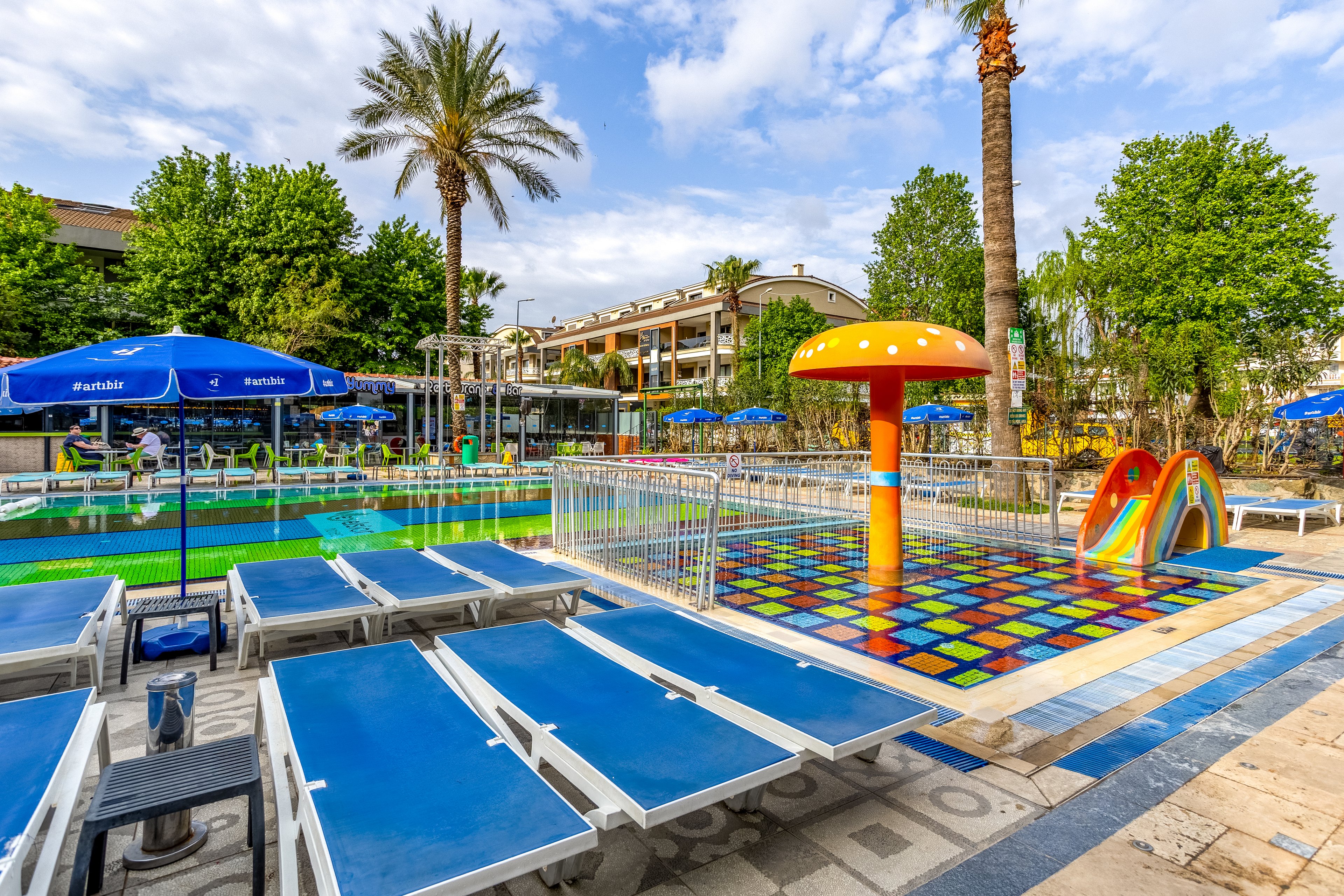 A vibrant children’s swimming pool area features a shallow pool with a colorful tiled floor and playful water structures. A large mushroom-shaped fountain and a small rainbow slide stand out as focal points. Blue sun loungers line the deck in the foreground, while blue umbrellas provide shade around the pool. Lush green trees and a multi-story resort building create a relaxed holiday atmosphere under a partly cloudy sky.