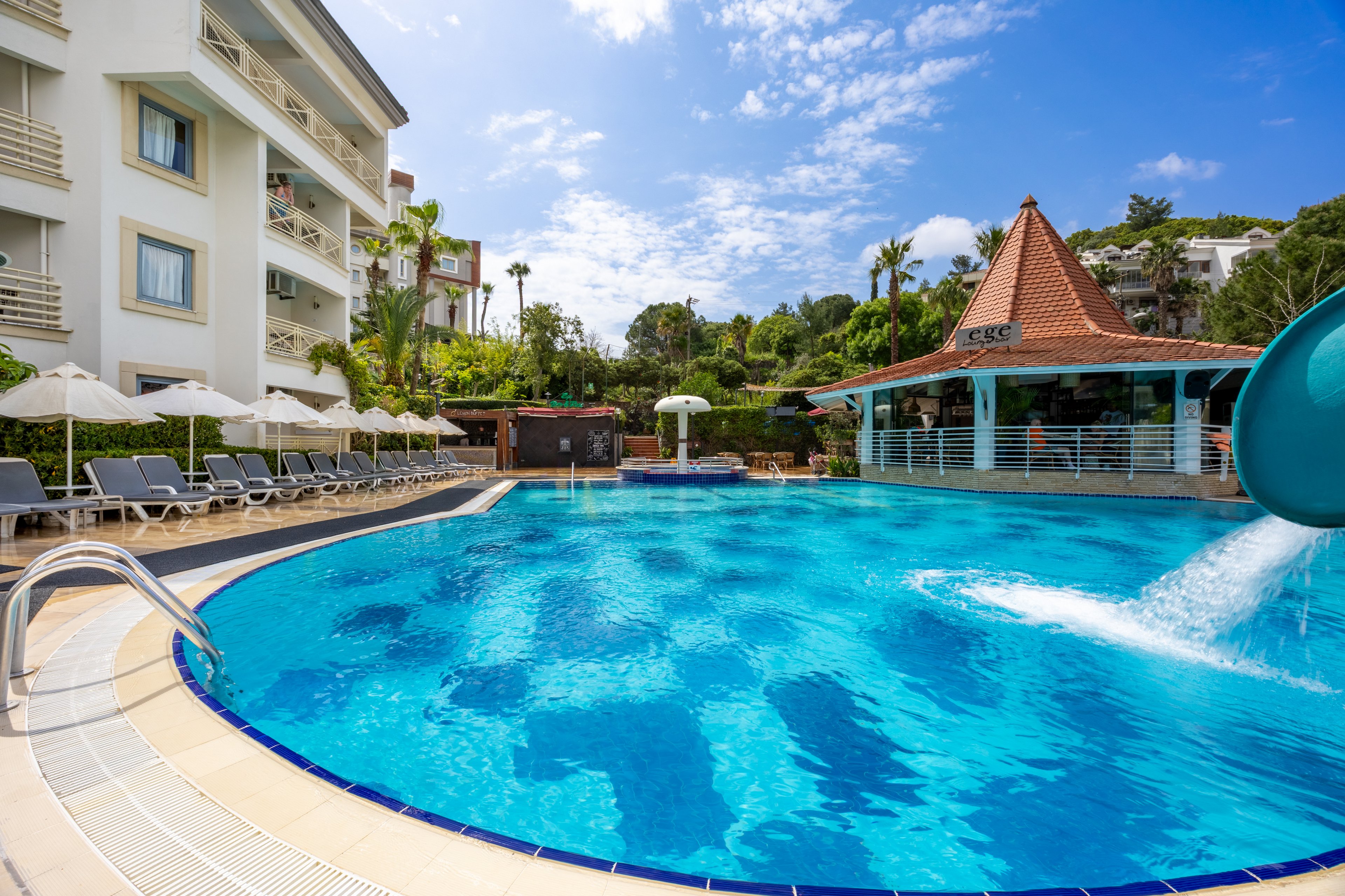 A large outdoor swimming pool is the main focus, surrounded by lounge chairs and umbrellas. The setting is a resort or hotel with a multi-story building and lush greenery in the background. A poolside bar with a red-tiled roof and a visible water slide add to the vacation atmosphere. The sky is bright and partly cloudy, enhancing the inviting mood. No visible text or numbers are present in the image.