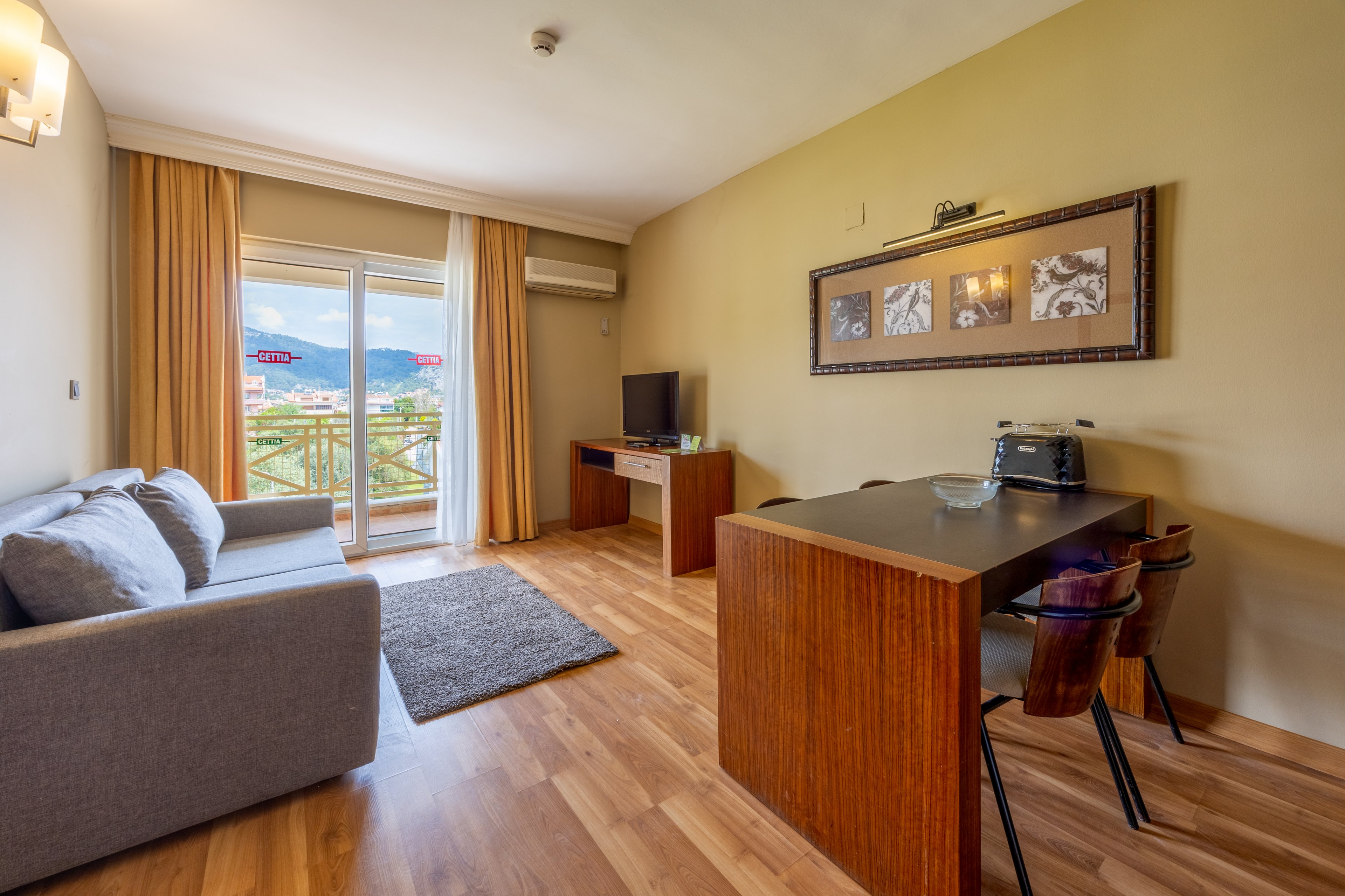 A contemporary hotel suite features a cozy gray sofa, wooden floors, and a compact dining area with two chairs. The room opens to a balcony with a scenic mountain view, visible through large glass doors. Neutral tones and natural light create a welcoming atmosphere, with a television and small appliances on the desk. Wall art and a soft rug add decorative touches to the space.