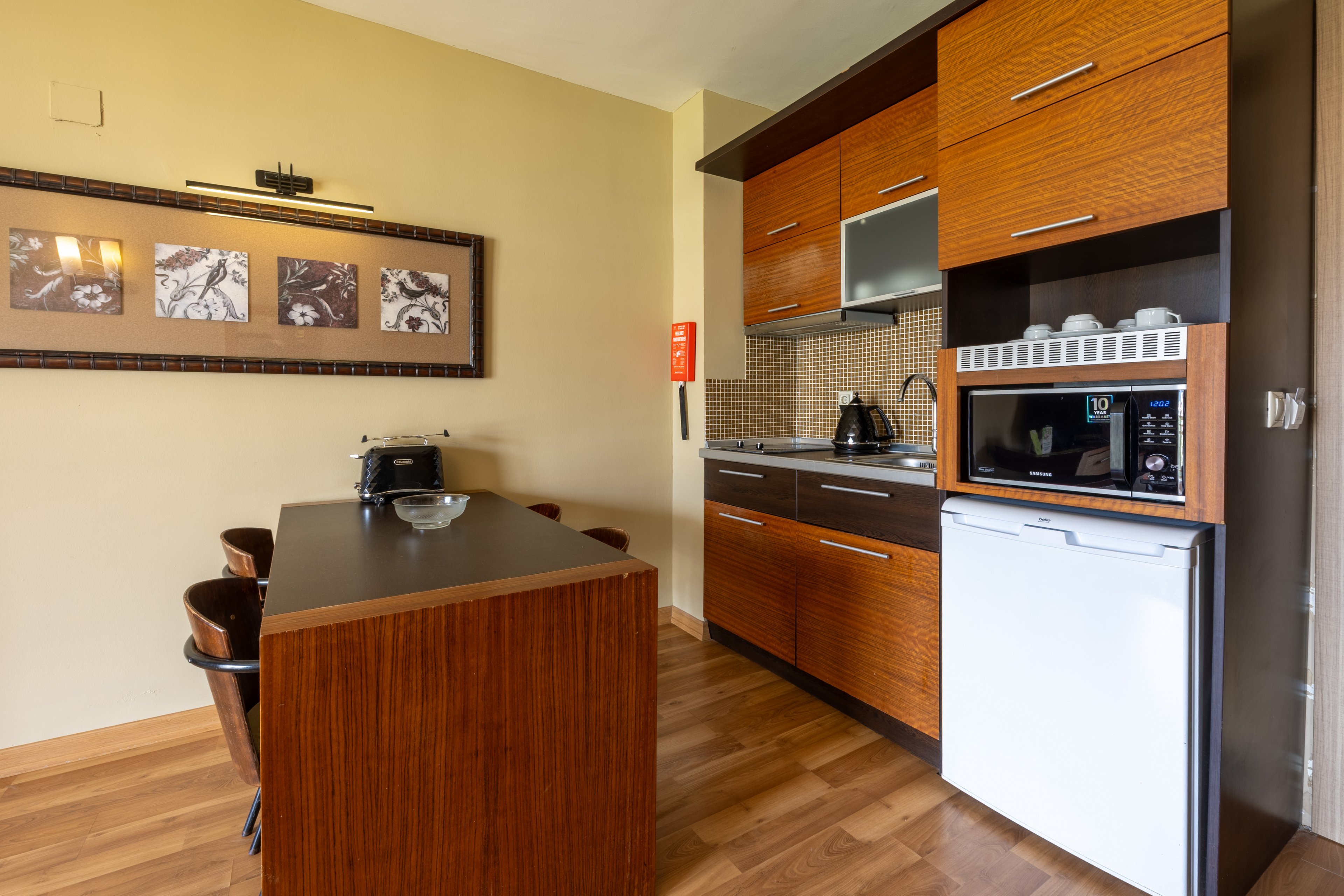 A contemporary kitchenette features wood cabinetry, a small refrigerator, microwave, and electric kettle. The setting includes a dark wood dining table with matching chairs, wall art, and a fire extinguisher mounted on the wall. The space is well-lit with a warm palette and clean, minimalist design. No visible people or explicit numeric values are present in the image.
