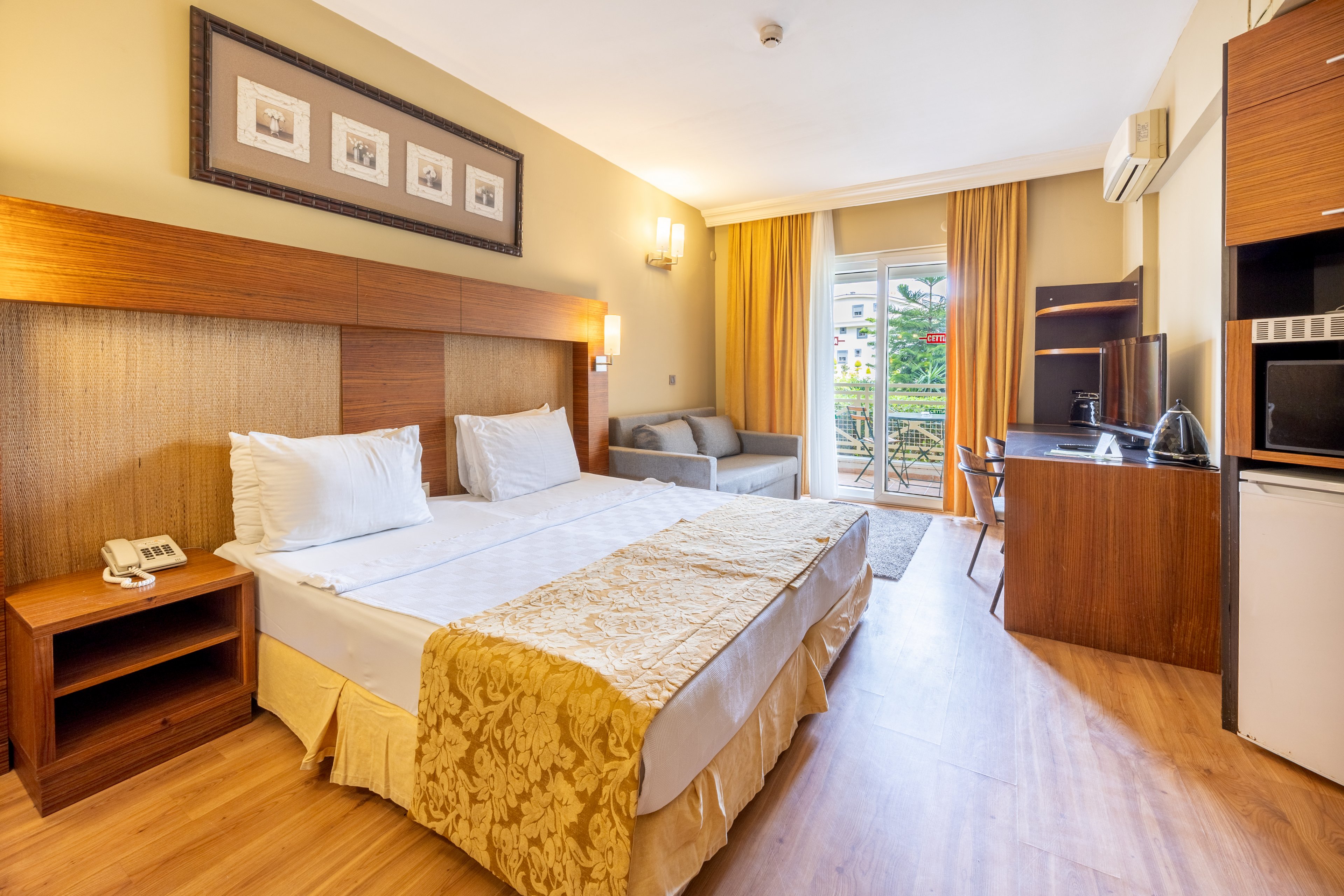 A spacious hotel room features a large double bed with white linens and a gold-patterned throw. The room includes a wooden desk, flat-screen TV, mini fridge, and a gray sofa near a glass door leading to a balcony with outdoor views. Warm lighting, framed wall art, and wood flooring create a welcoming, contemporary atmosphere.