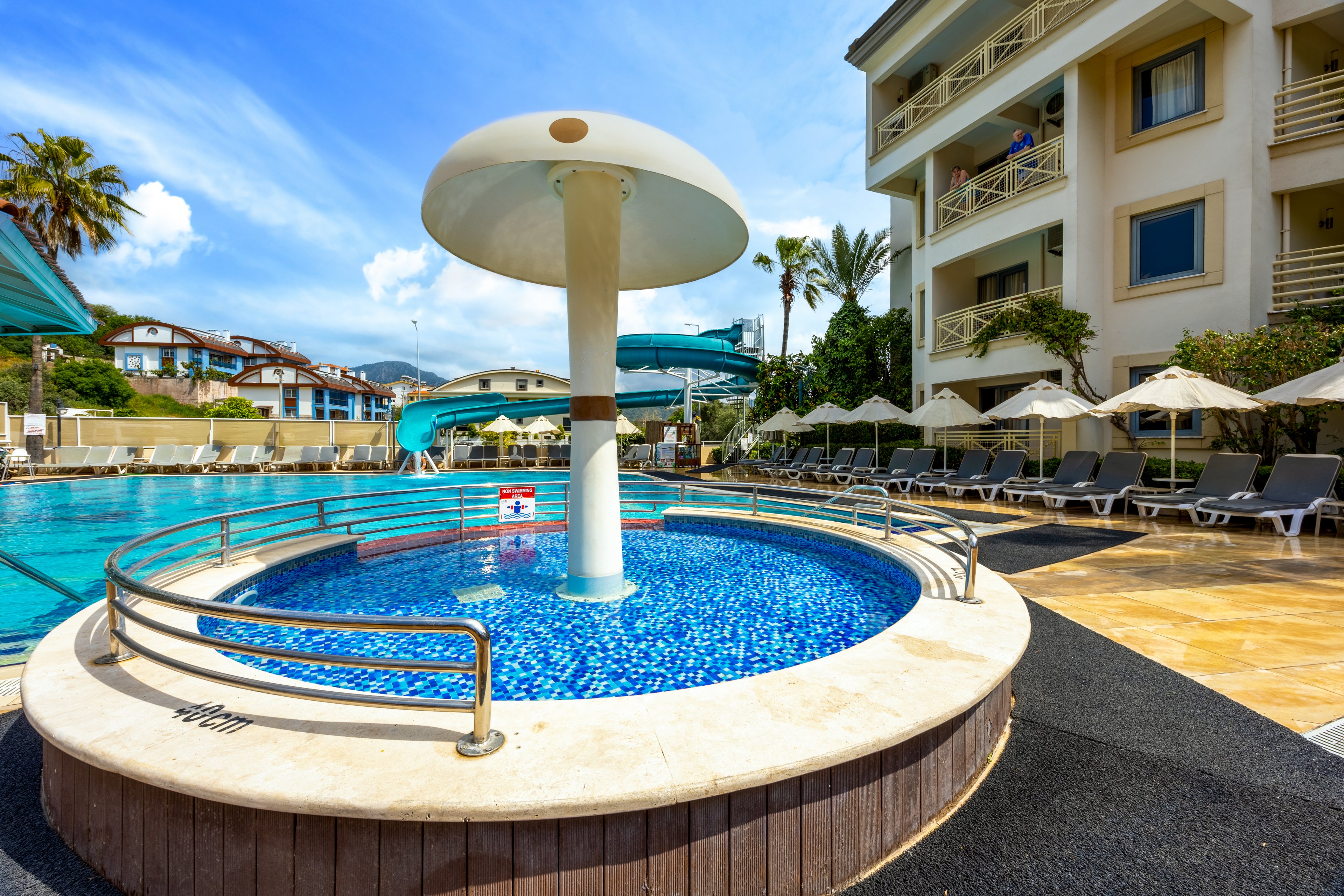 A vibrant outdoor resort pool area features a circular children's pool with a large mushroom-shaped water fountain at its center. Surrounding the pool are lounge chairs under umbrellas, a waterslide, and a multi-story hotel building. The setting is bright and inviting, with clear blue skies and lush palm trees enhancing the vacation atmosphere. No visible text or numbers are present in the image.
