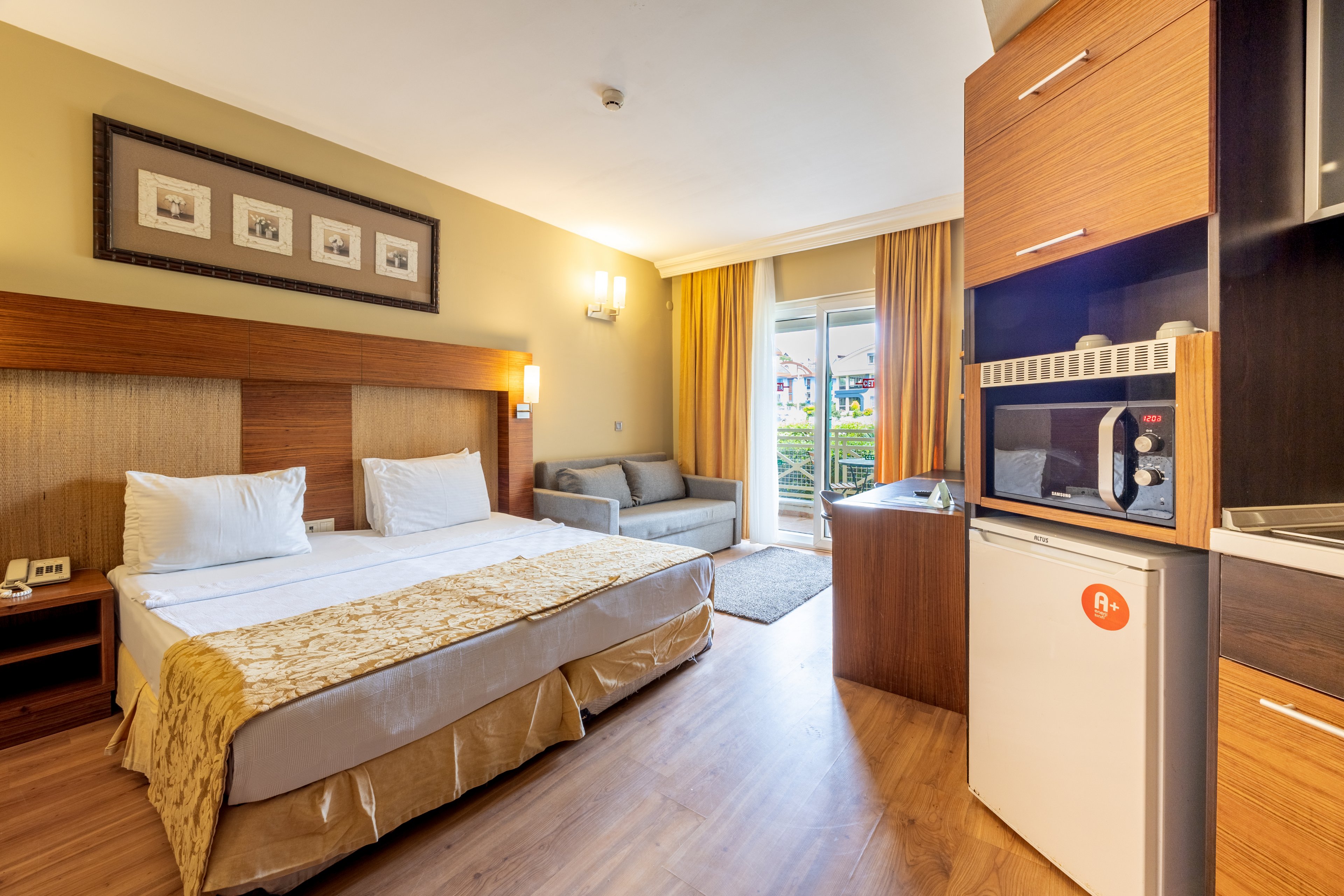 A spacious hotel room features a double bed with white linens and a gold runner, set against a wooden headboard. The room includes a small kitchenette area with a microwave and mini fridge, as well as a cozy seating area with a gray sofa near a balcony door. Warm lighting, wood flooring, and neutral tones create a welcoming atmosphere, while framed artwork and curtains add decorative touches.