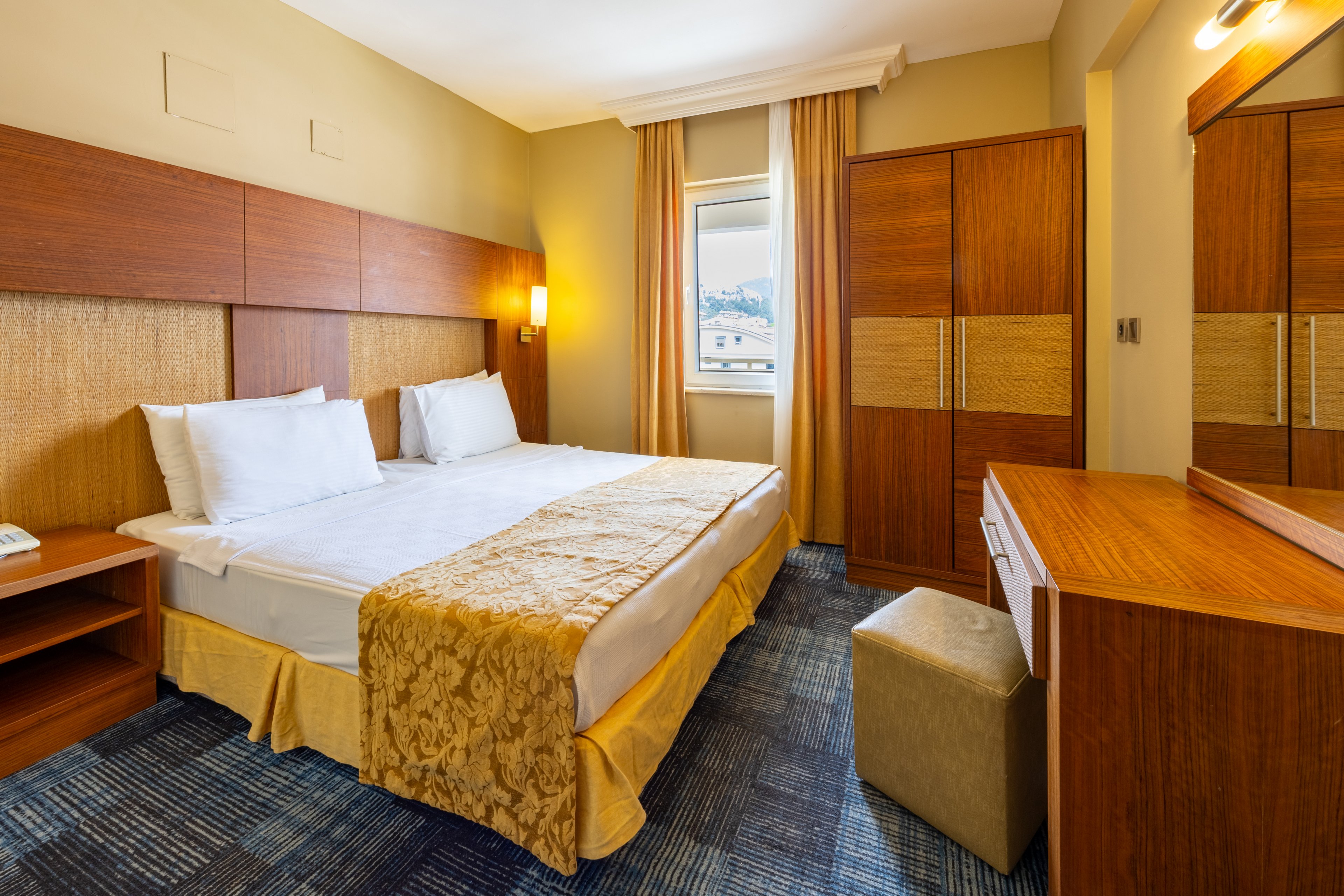A contemporary hotel room features a neatly made double bed with white linens and a gold-patterned runner. The space includes wooden furniture such as a wardrobe, desk, and bedside tables, with a window allowing natural light and a view outside. Warm yellow and beige tones create a cozy, inviting atmosphere. The room is well-lit with wall-mounted lamps and has a blue-patterned carpet. No visible text or numbers are present in the image.