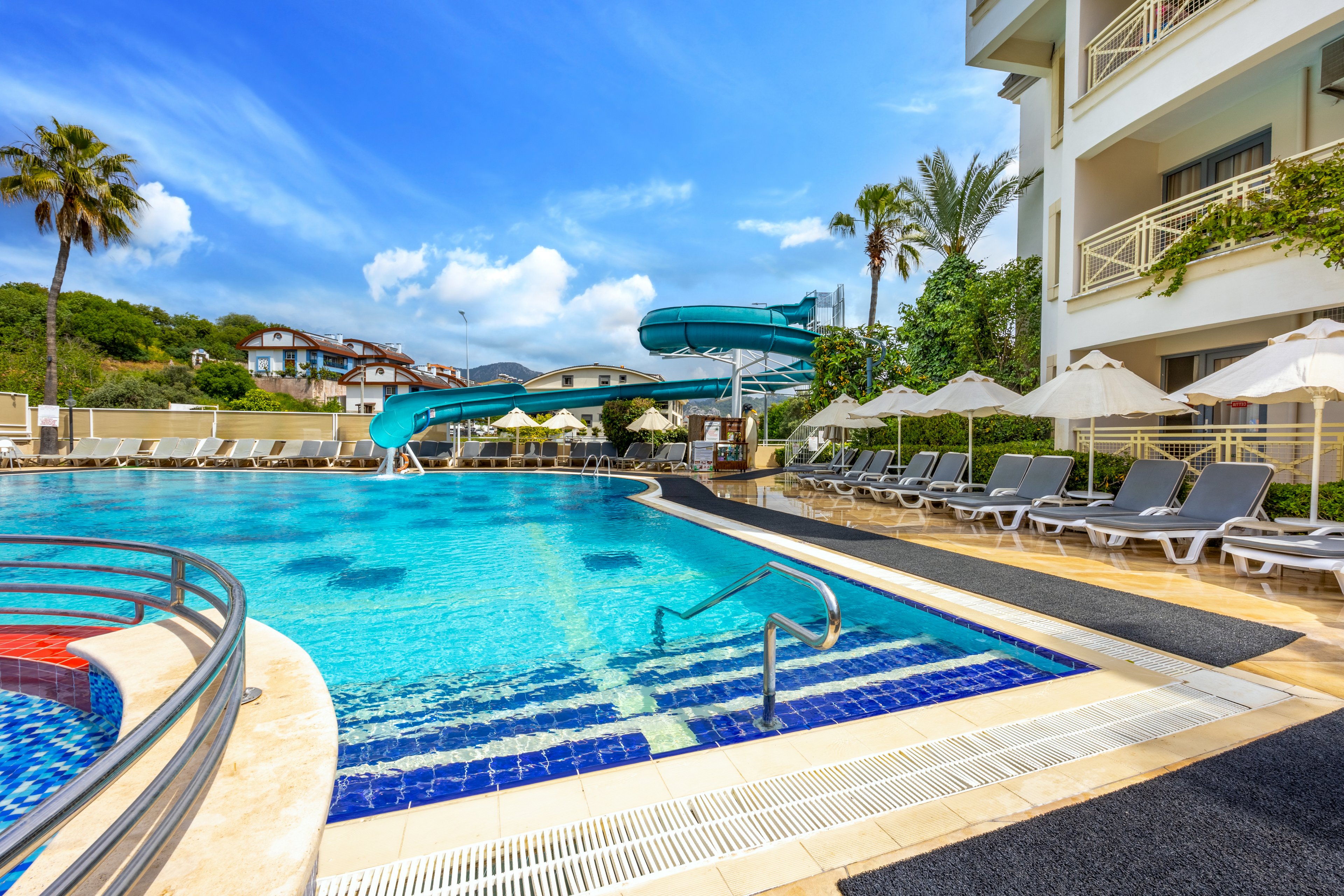 A large outdoor swimming pool is featured at a modern resort, with a blue waterslide curving into the water. The poolside area is lined with numerous gray lounge chairs and white umbrellas, creating a relaxing atmosphere. The setting is bright and sunny, with palm trees and landscaped greenery in the background. The building’s balconies overlook the pool, enhancing the vacation vibe. No visible text or numbers are present in the image.
