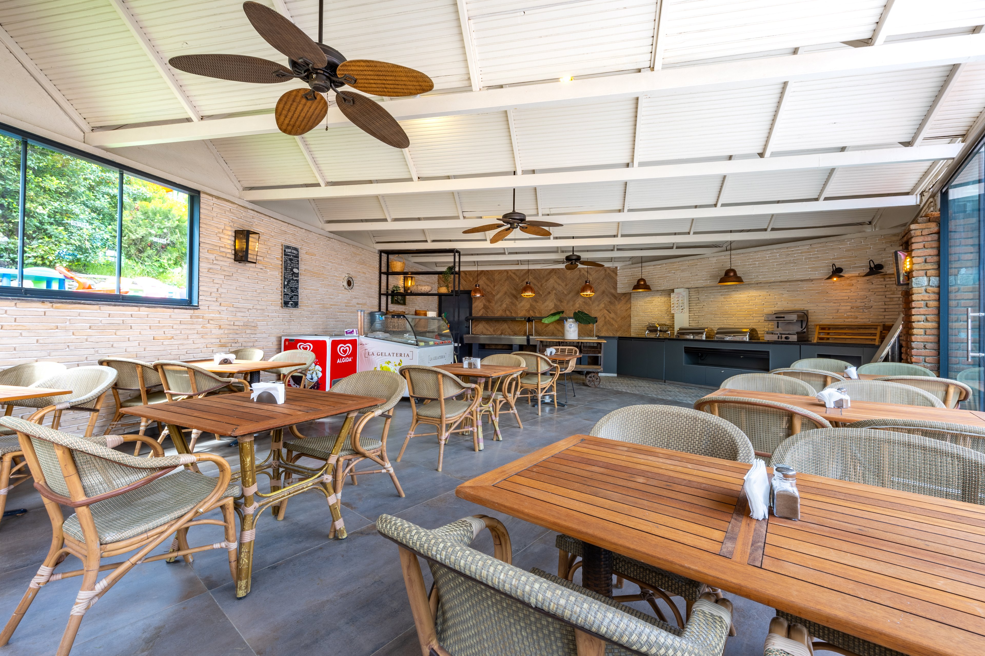 A spacious café interior features wooden tables and wicker chairs arranged neatly under a white ceiling with ceiling fans. The setting is airy and well-lit, with large windows revealing greenery outside. A counter area and visible beverage coolers, including a clearly marked 'Coca-Cola' cooler, are present. The overall mood is relaxed and inviting, with neutral tones and natural materials. No people are present in the scene.